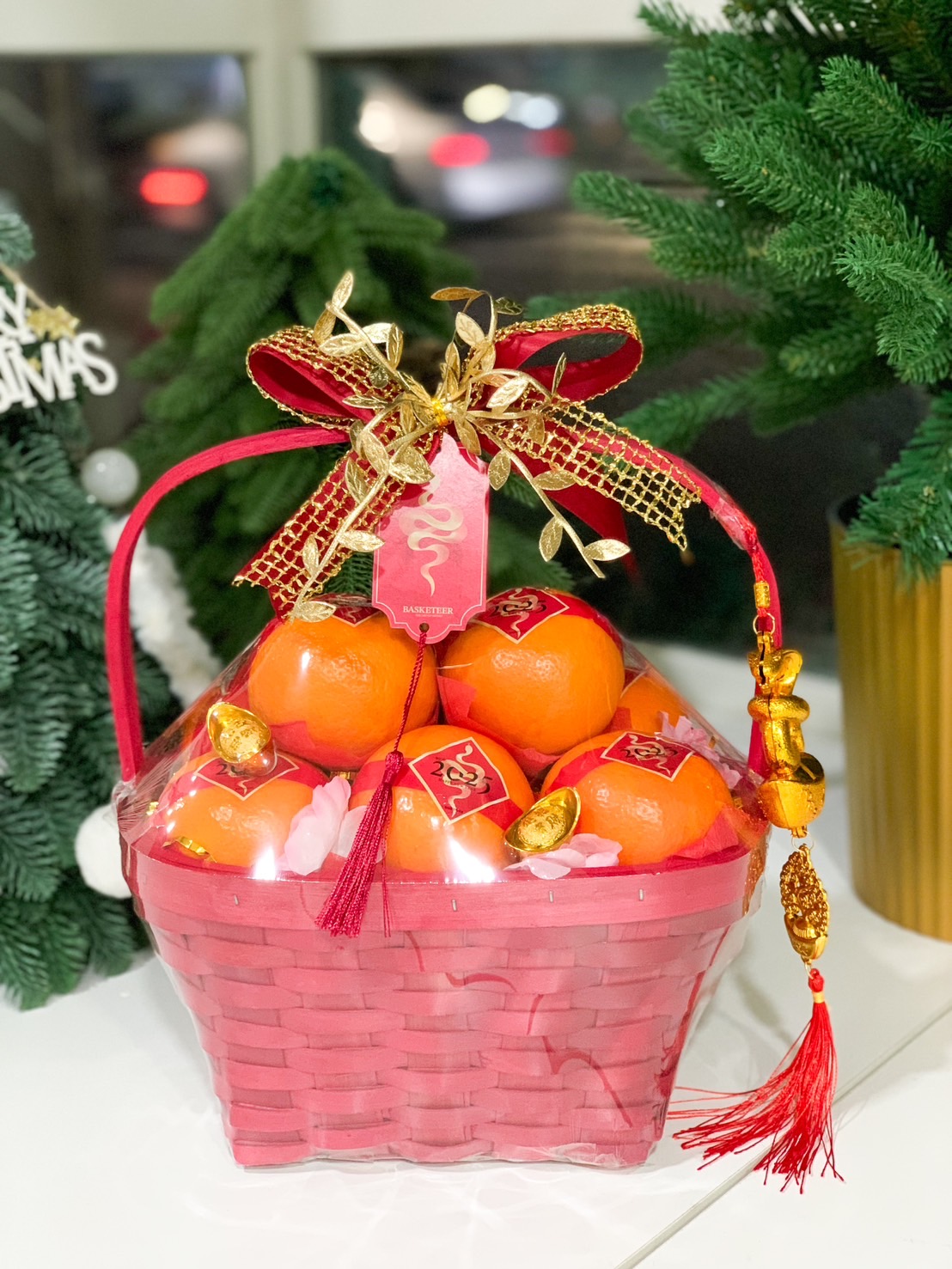 Red gift baskets filled with ripe persimmons wrapped in clear plastic. Each basket is decorated with pink flowers and adorned with decorative red bows and golden ornaments. The baskets are placed on a white surface against a plain white background.