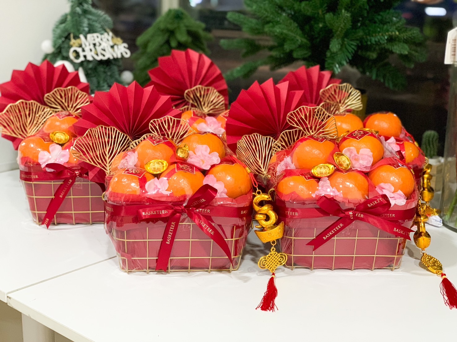 Beautifully designed Chinese New Year hampers featuring fresh oranges, gold wire baskets, red festive fans, pink blossoms, and golden charms, perfect for gifting during the celebrations.