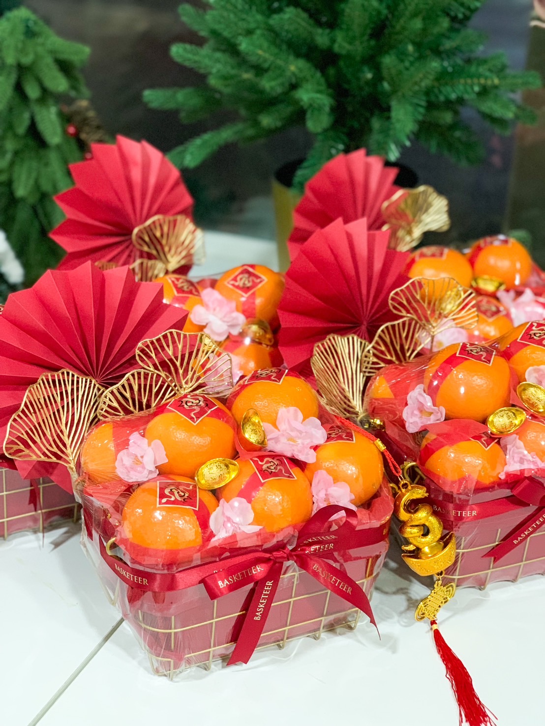 Close-up of a Beautifully designed Chinese New Year hampers featuring fresh oranges, gold wire baskets, red festive fans, pink blossoms, and golden charms, perfect for gifting during the celebrations.