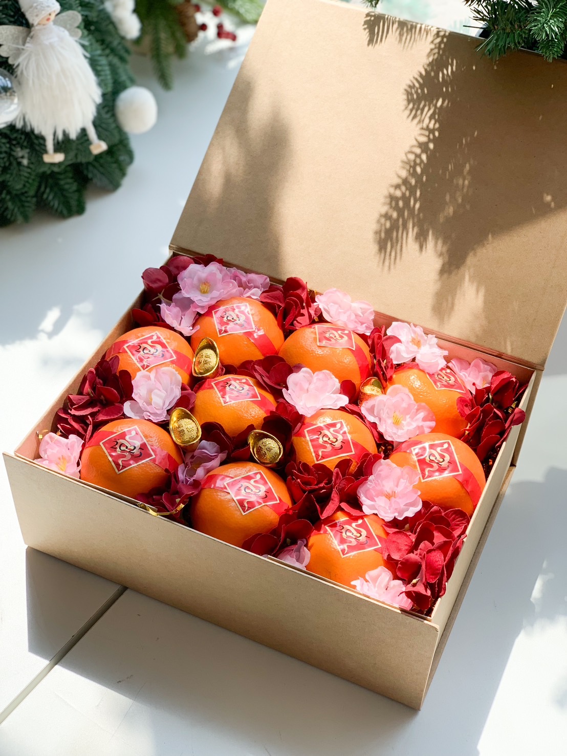 Mandarin oranges neatly arranged in a box, adorned with red and pink flowers, gold ingots, and festive Chinese New Year decorations.