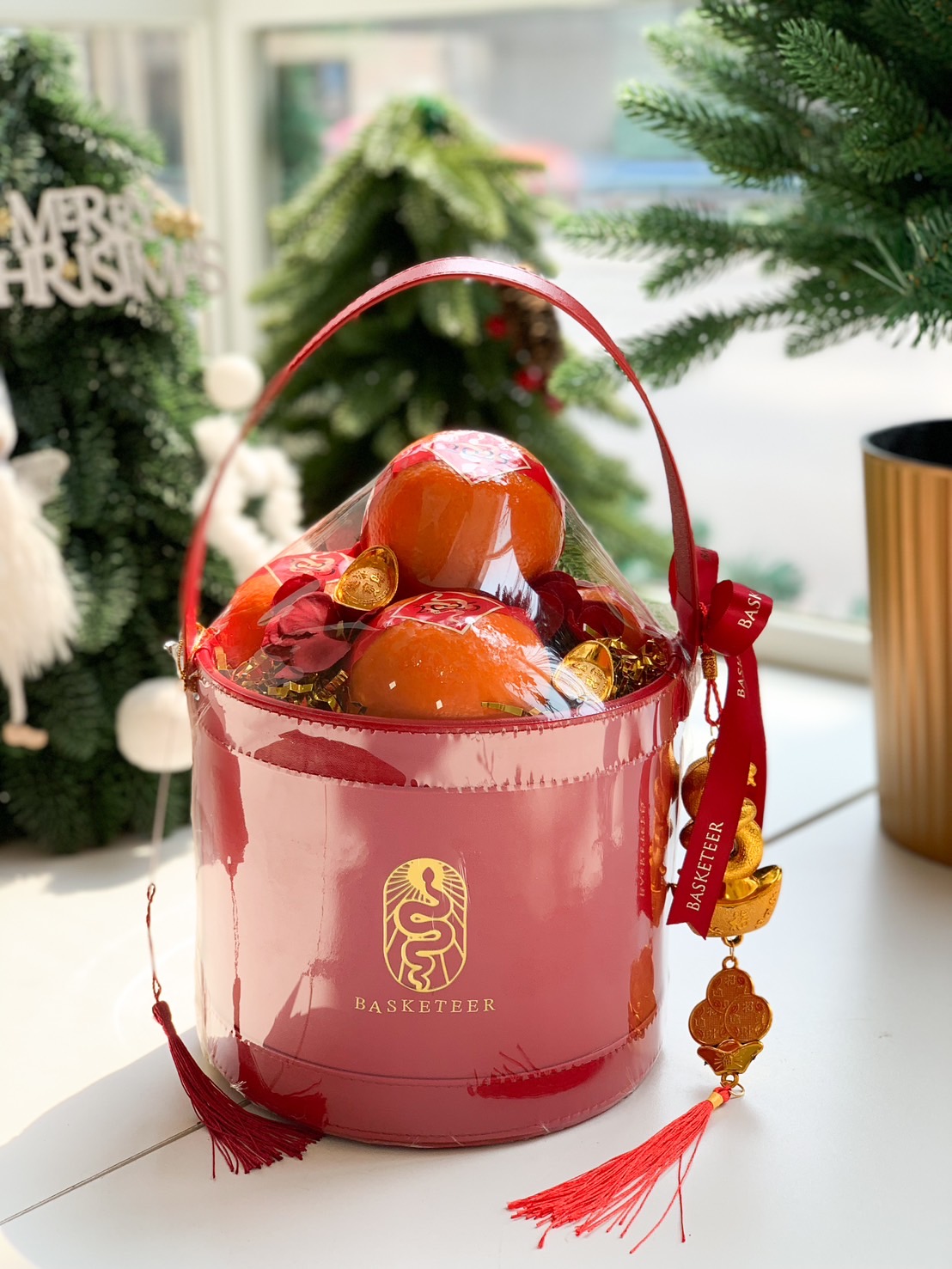 A round red basket with a red handle and bow, filled with oranges and a decorative gold charm. The basket features a yellow Chinese character on the front and is placed on a white surface. The background is plain white.