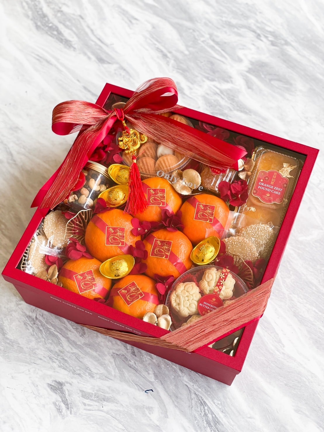 A festive red gift box adorned with a ribbon and gold charm, containing an assortment of treats including oranges, gold foil-wrapped chocolate coins, cookies, and assorted snacks. The box has a clear lid showcasing the contents. The background is a marble surface.