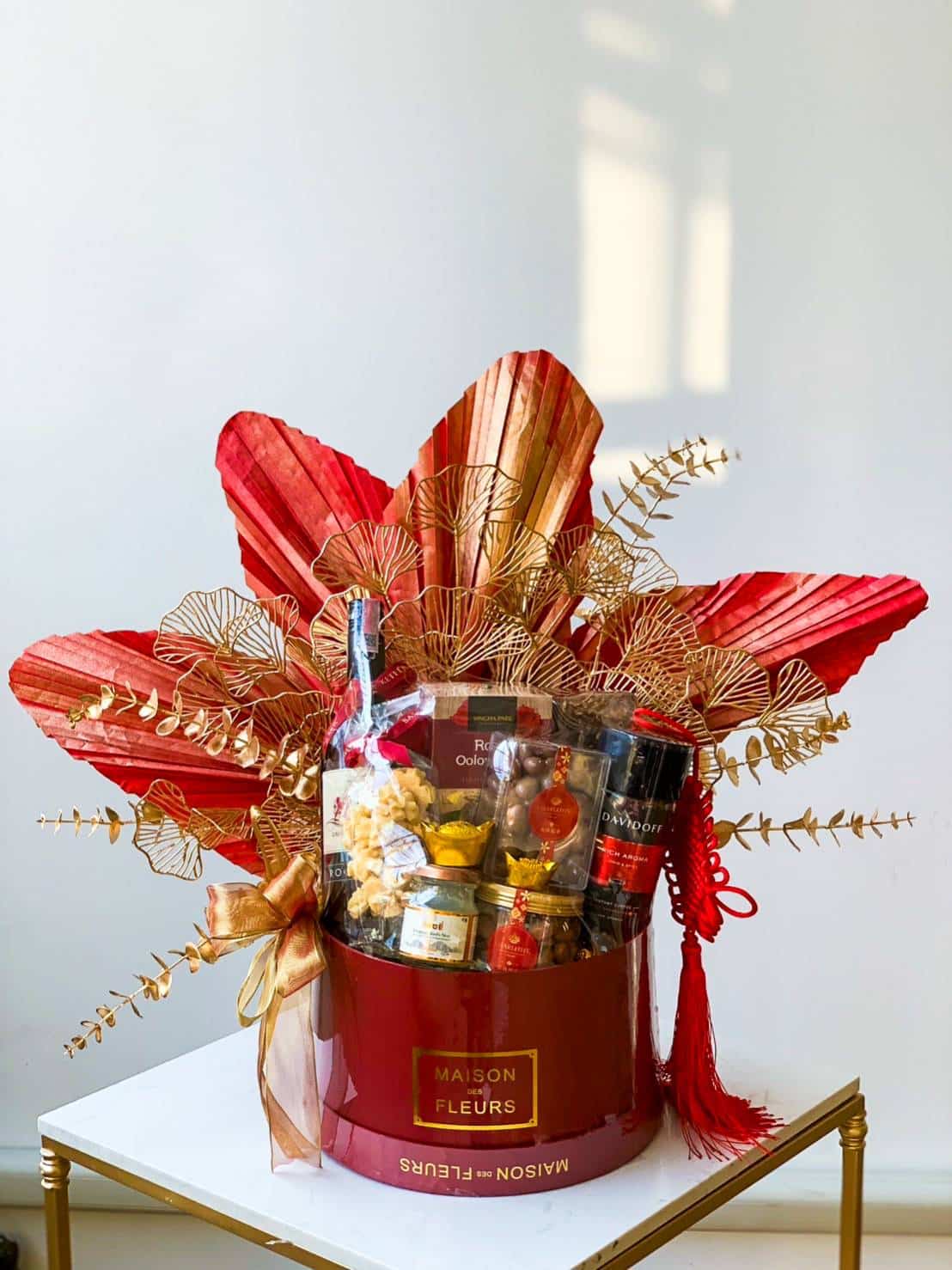 A red and gold themed gift basket featuring folded and decorative red paper fans, gold leaves, and ribbon on top. The basket contains various food items such as chocolates, snacks, and a bottle, all elegantly arranged within a red container labeled 
