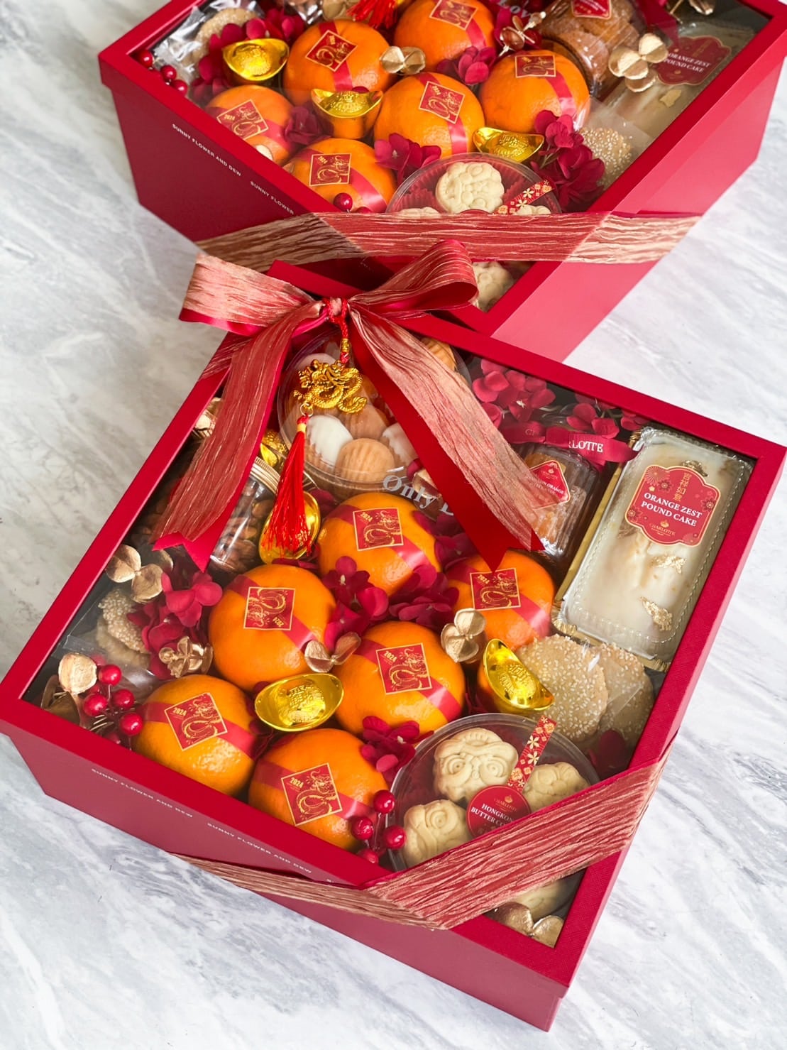 Two red gift boxes are filled with a variety of festive treats, including ornamental items, snacks, and persimmons. Each box is topped with a decorative red and gold ribbon, adding a luxurious touch. The background is a marble surface.