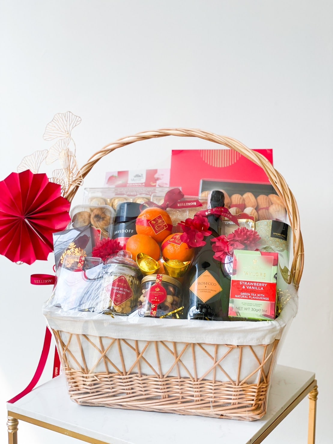 A wicker gift basket filled with an assortment of items including wine, chocolate, citrus fruits, pastries, and other treats. The basket is decorated with red accents and a red ribbon bow, placed against a plain white background.