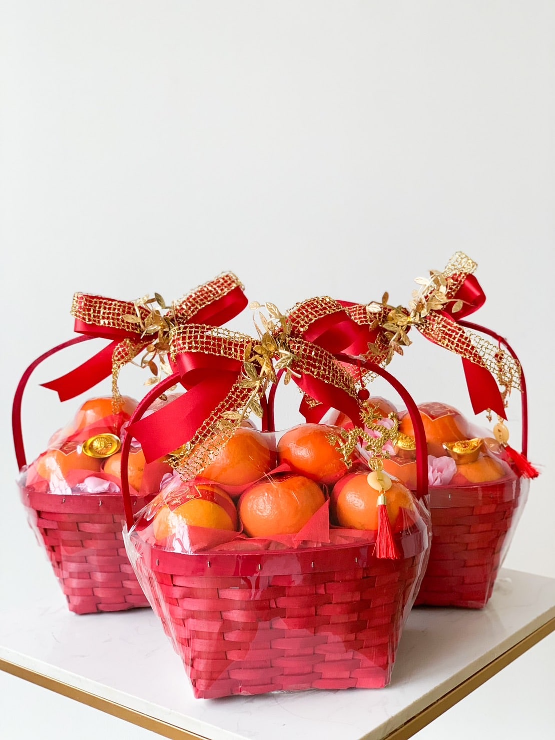 Three red wicker baskets, each filled with oranges and wrapped with decorative red and gold ribbons. The baskets also contain small gold items and are adorned with gold trinkets and charms. They rest on a white surface against a plain background.