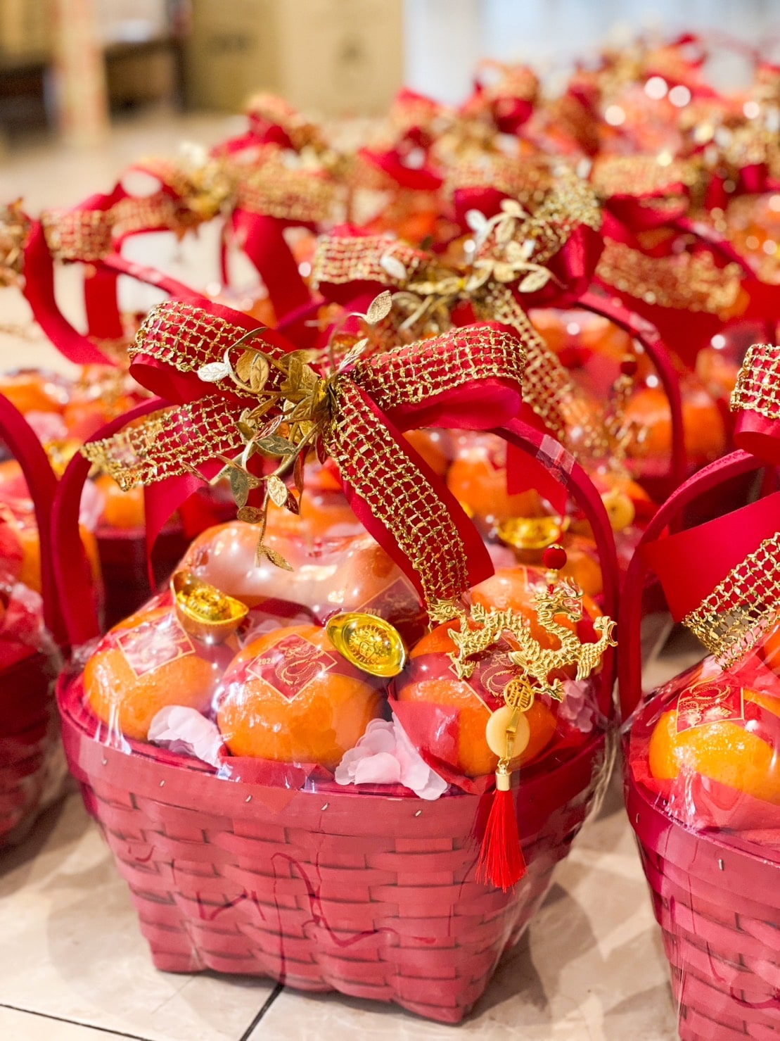 A collection of decorative red baskets filled with oranges and sweets, each adorned with a large red ribbon and gold ornaments. The baskets are arranged in rows for a festive occasion, likely celebrating Chinese New Year.