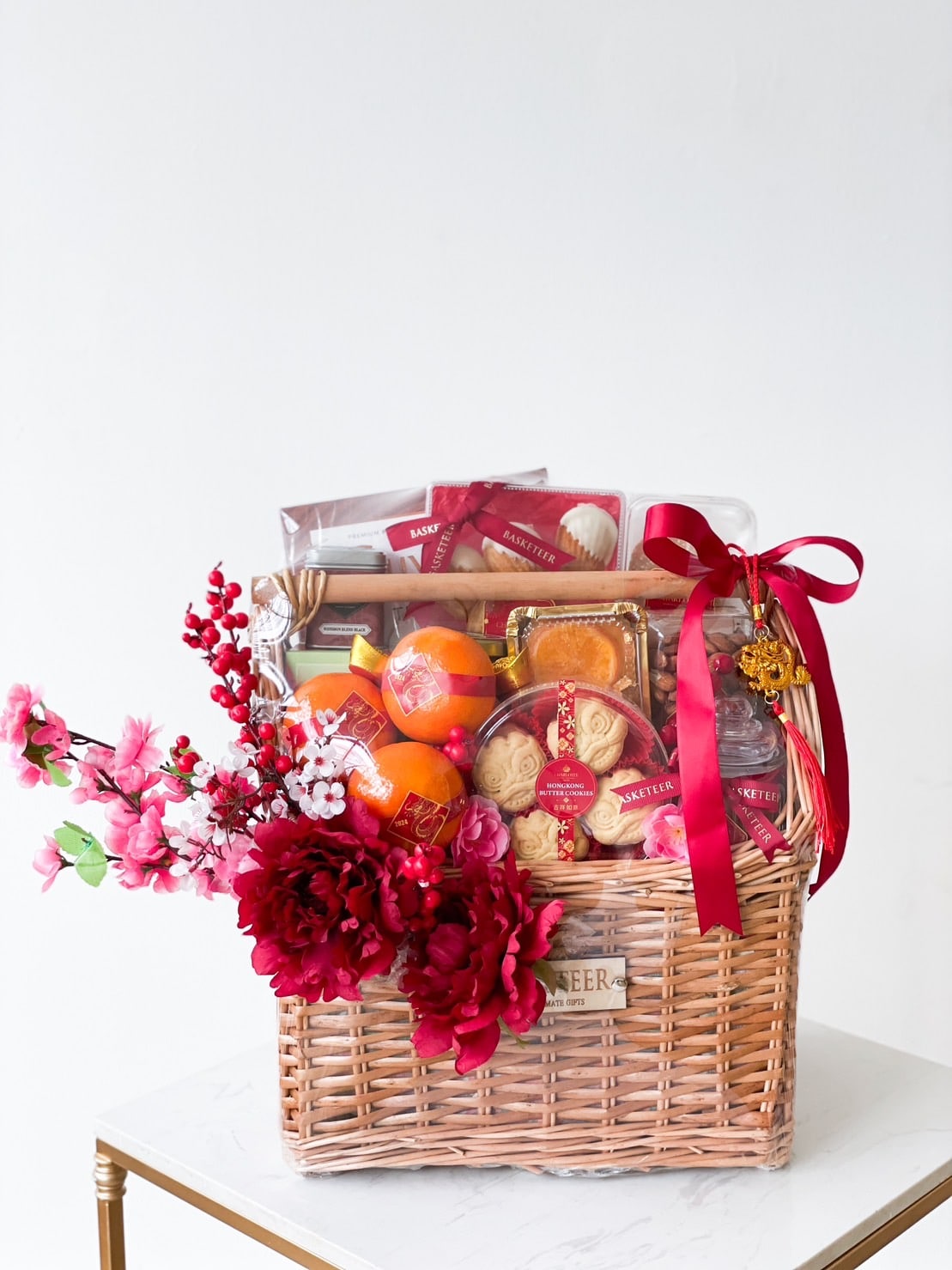 A gift basket filled with various packaged snacks, fruits, and cookies. The wicker basket is adorned with a red ribbon and decorated with red and pink artificial flowers. The basket sits on a white marble surface with a plain white background.