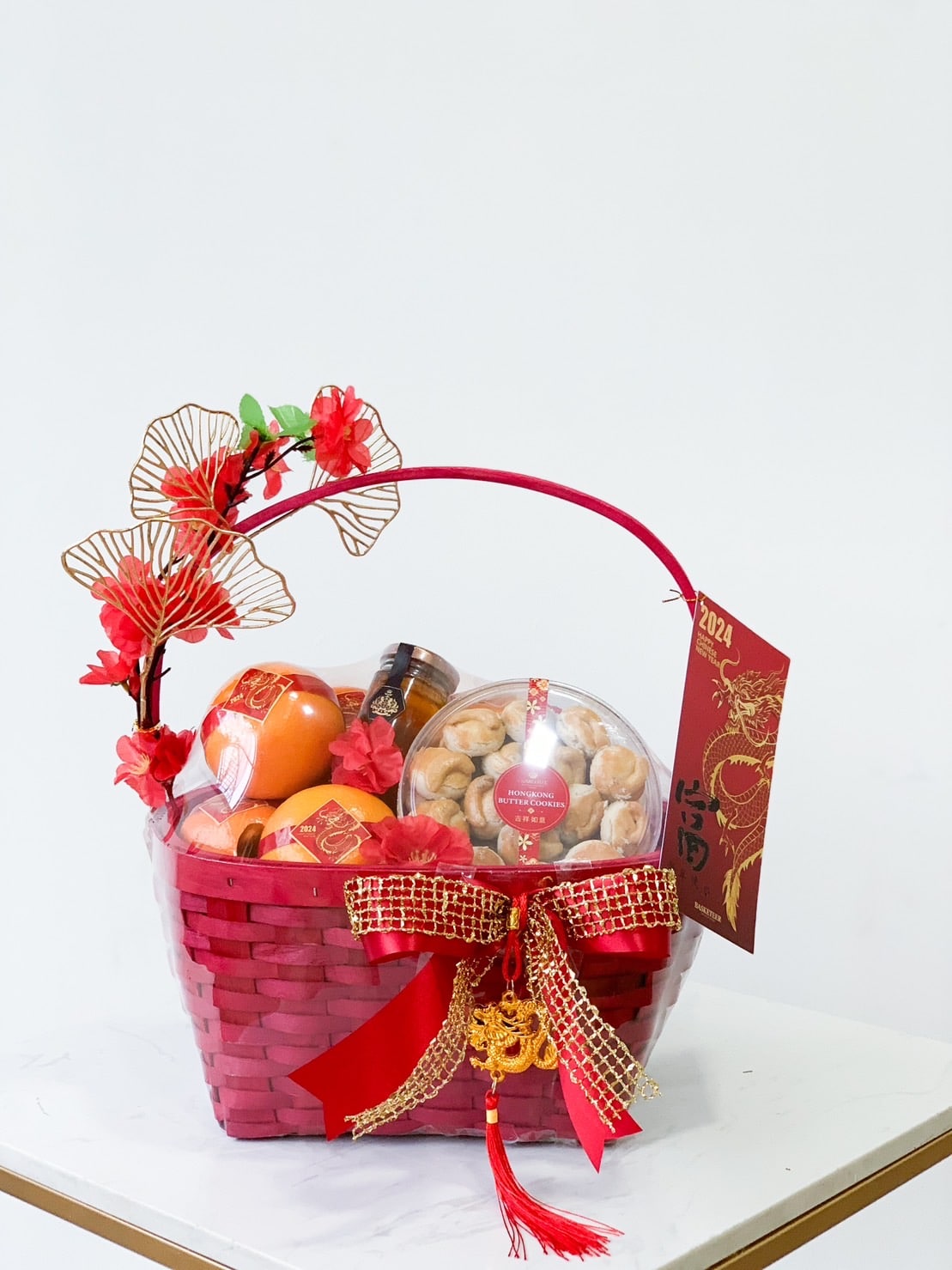 A red woven gift basket adorned with gold and red decorations, including flowers and a bow, contains a variety of packaged treats. A red envelope with a golden dragon design and the year 