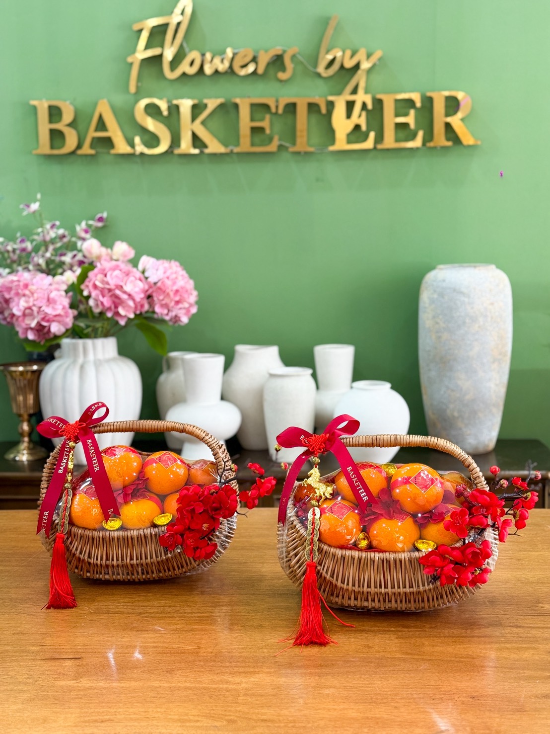 Two wicker Festive Mandarin Orange Carryall Gift Hampers, decorated with red ribbons and tassels, sit on a wooden table. Behind them are white vases, pink flowers, and a green wall with the gold sign 