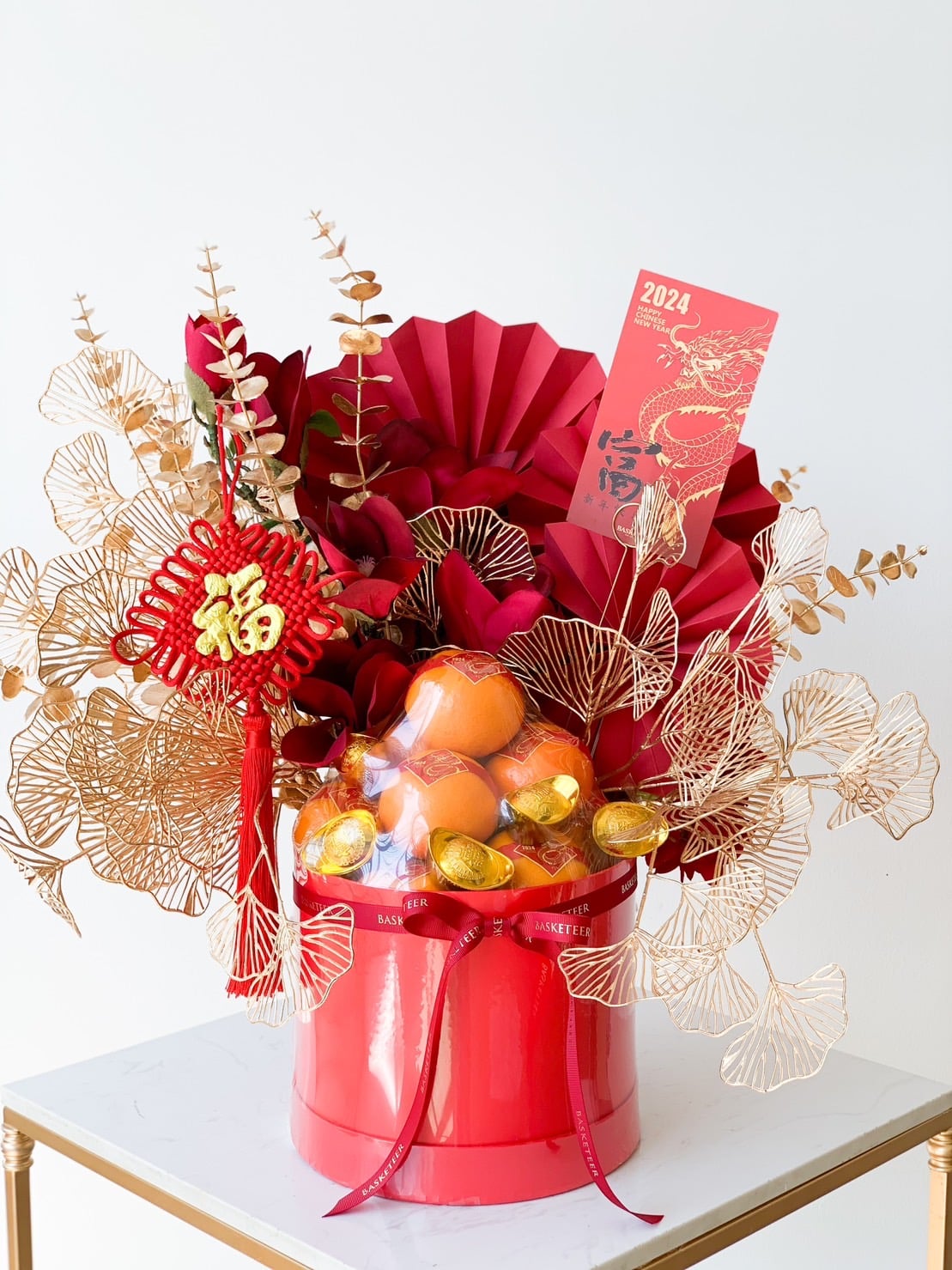 A vibrant arrangement for Lunar New Year featuring red and gold elements. The centerpiece includes gold foil-wrapped chocolates, oranges, red flowers, gilded leaves, and a red envelope with a dragon design and the year 2024. A red knot charm with a Chinese character is attached.