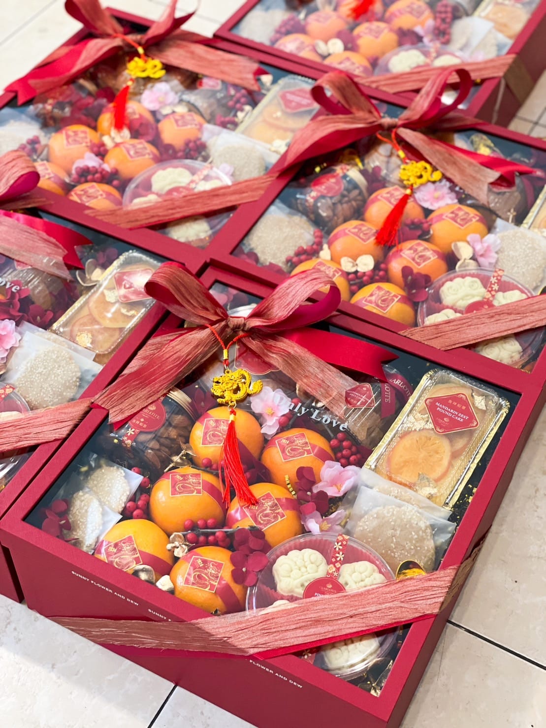 Gift boxes adorned with red ribbons containing oranges, golden packets, pastries, and other festive treats are arranged neatly. Decorative charms featuring gold symbols and flowers add a celebratory touch, conveying a festive and joyful atmosphere.