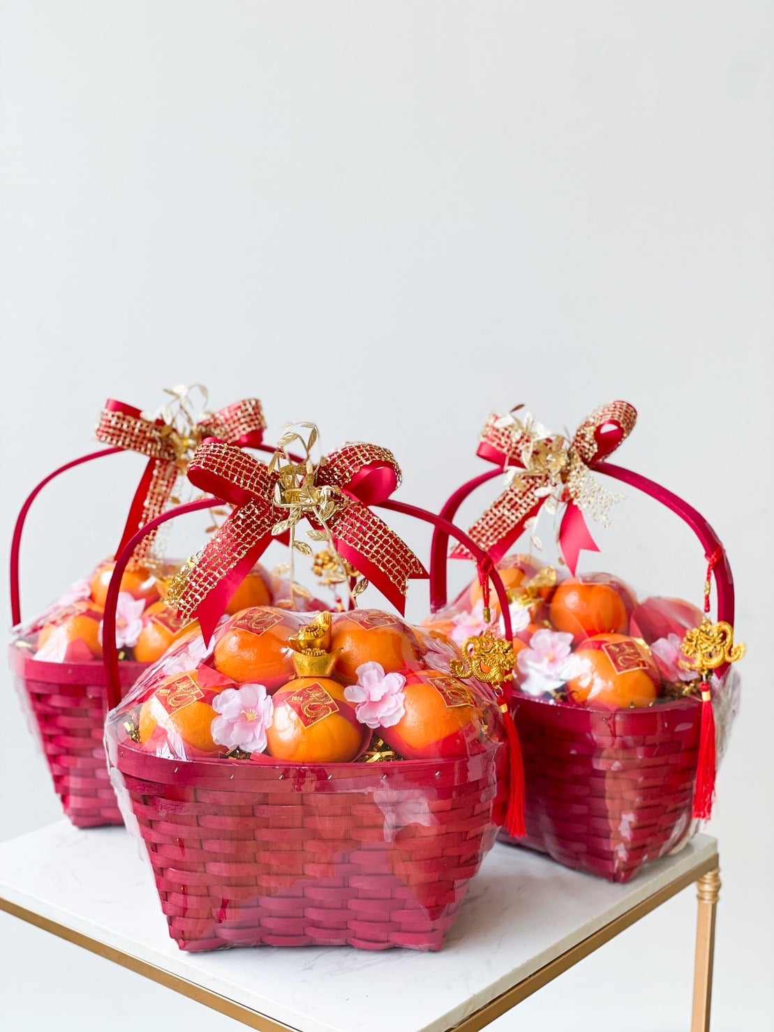Three red gift baskets filled with ripe persimmons wrapped in clear plastic. Each basket is decorated with pink flowers and adorned with decorative red bows and golden ornaments. The baskets are placed on a white surface against a plain white background.