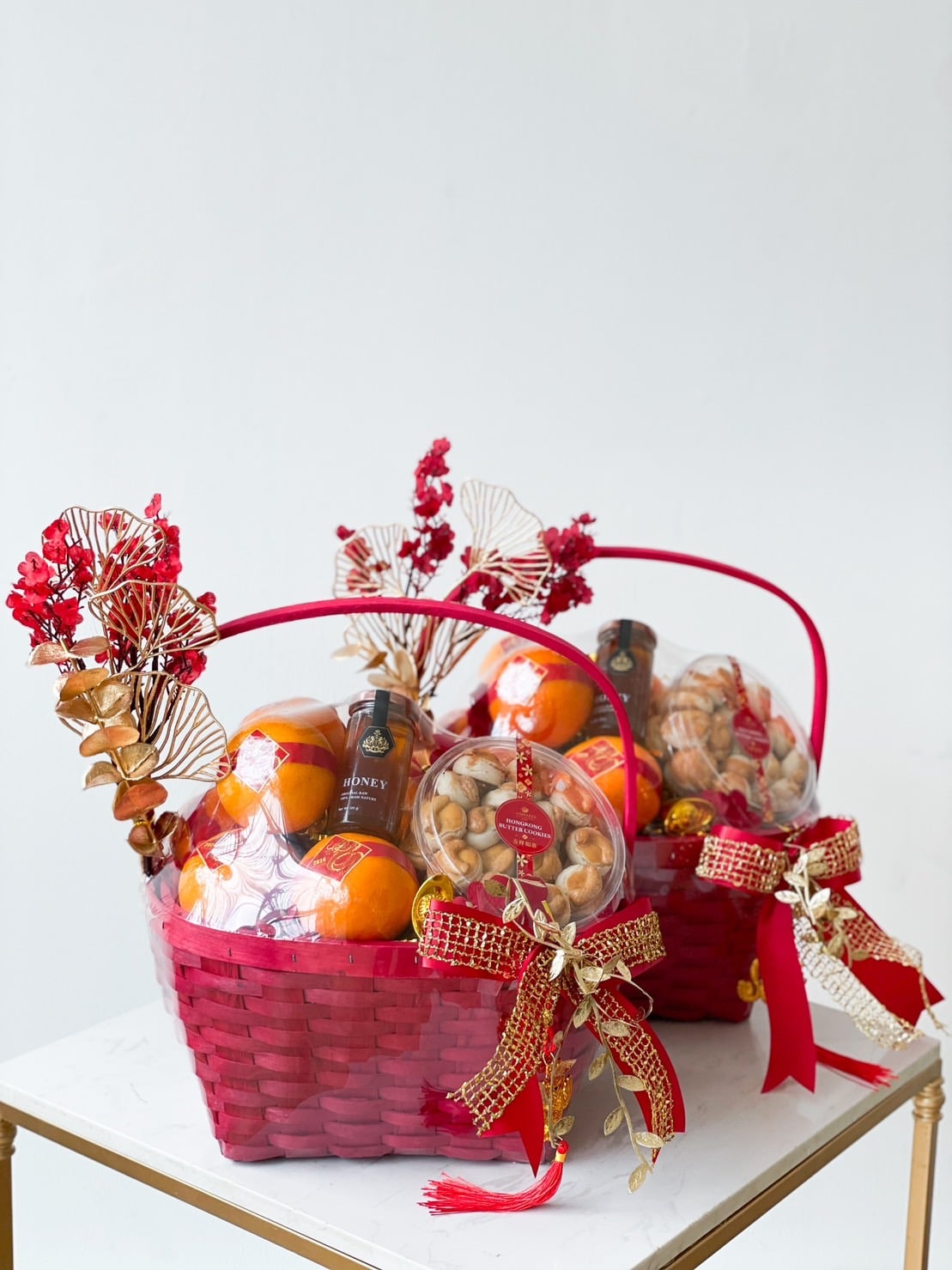 Two decorative red gift baskets are filled with oranges, a bottle of honey, a container of nuts, and dried flowers. Each basket is adorned with a large red and gold bow, along with fan-shaped embellishments and small red flowers.