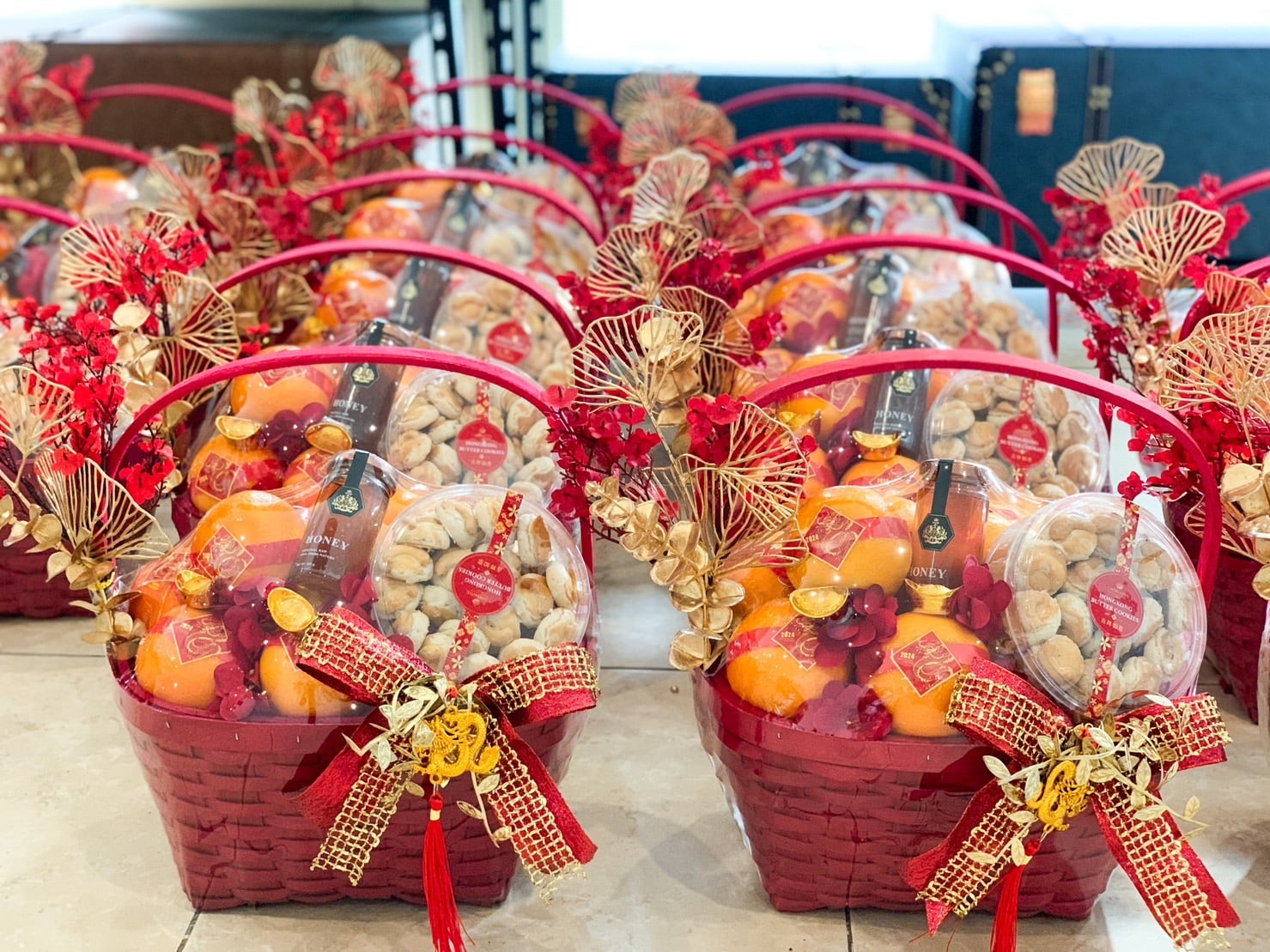 Red wicker baskets arranged in rows, each filled with an assortment of fresh oranges, packaged snacks, and decorated with red and gold ribbons, flowers, and festive golden ornaments. The baskets are placed on a tiled surface.