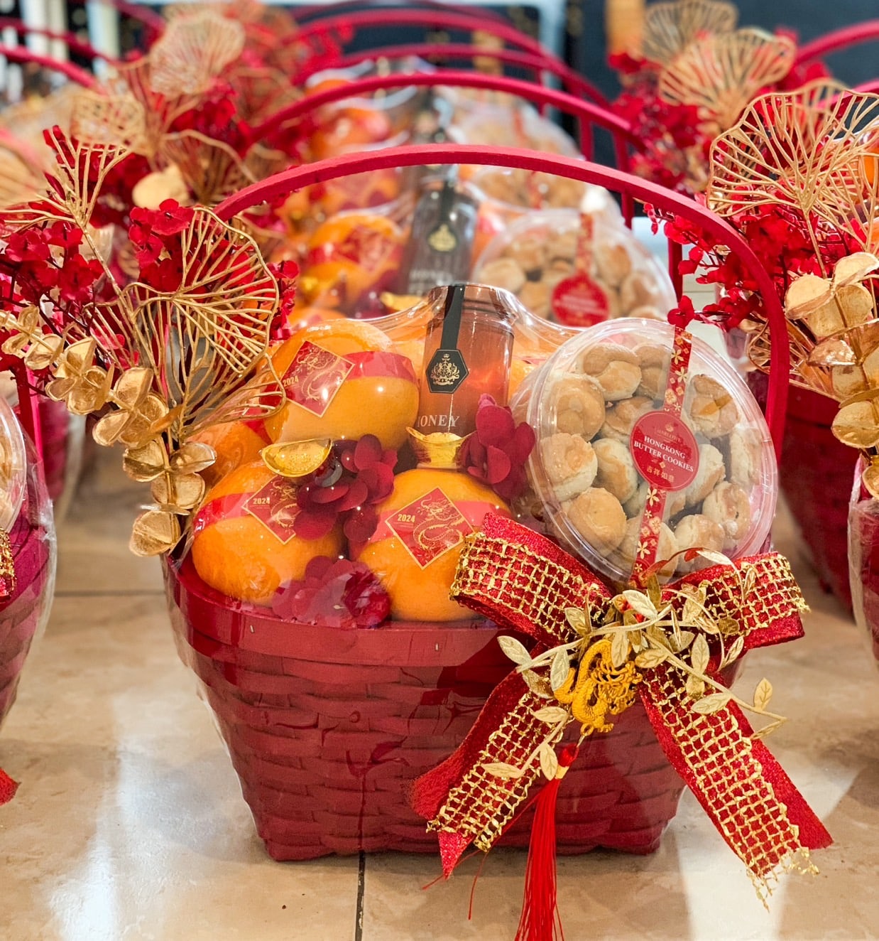A beautifully arranged red gift basket containing oranges, a box of cookies, and a bottle of honey. The basket is adorned with golden and red decorative elements, including flowers and a ribbon. Other similar baskets are visible in the background.