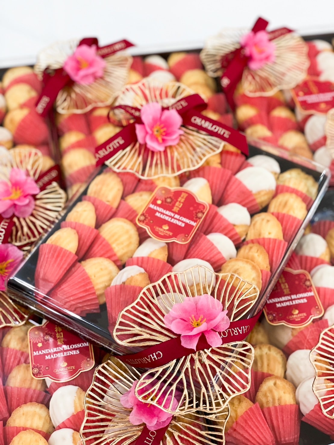 A display of gift boxes containing assorted madeleine cookies, wrapped in transparent lids adorned with red bows and decorative flowers. The cookies are arranged in alternating patterns and labeled 