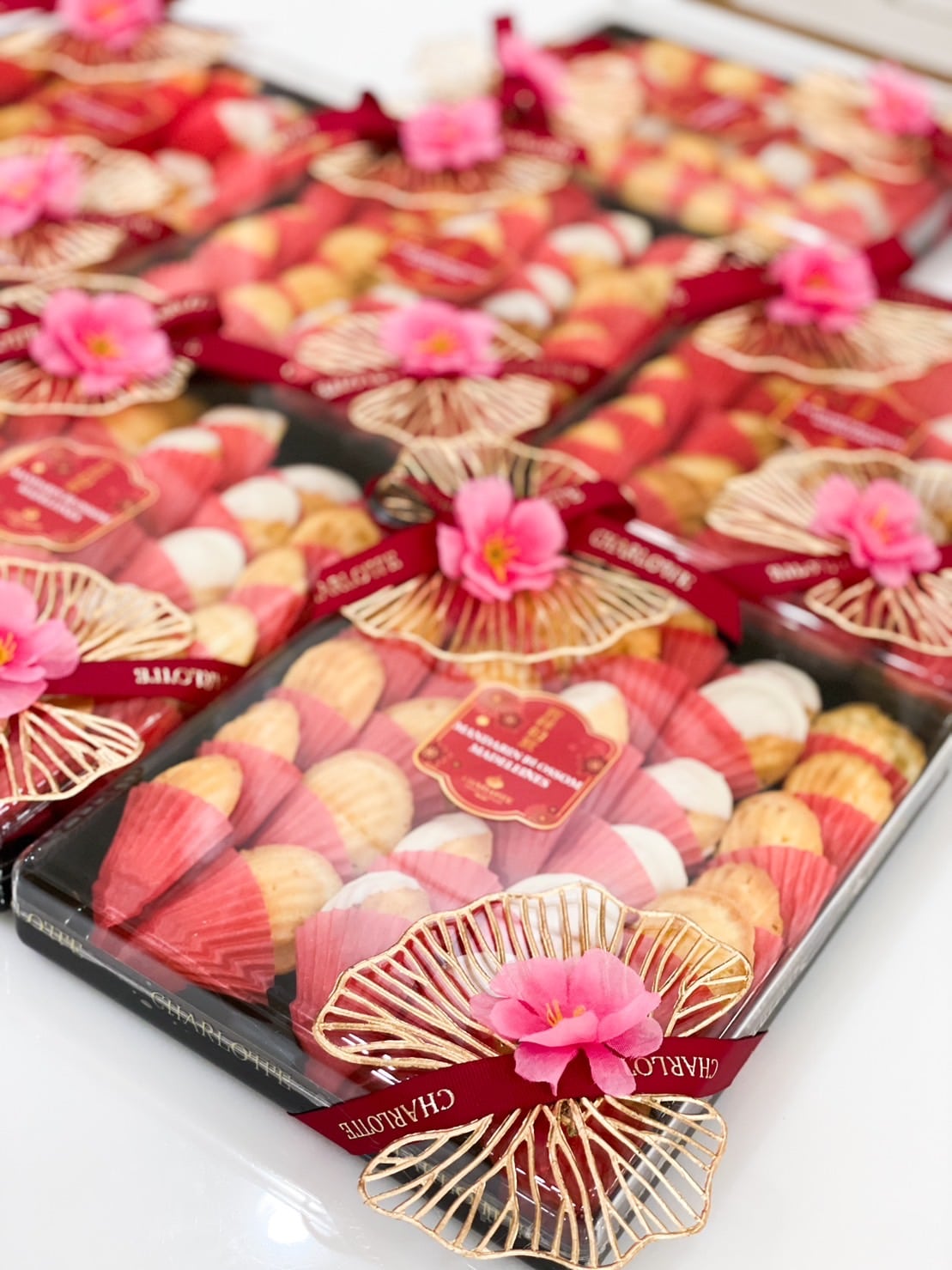 Boxes of assorted pastries wrapped in colorful liners are neatly arranged. Each box is adorned with a red ribbon and an intricate golden bow, topped with a pink artificial flower. A decorative label can be seen in the center of each box. The backdrop is white.