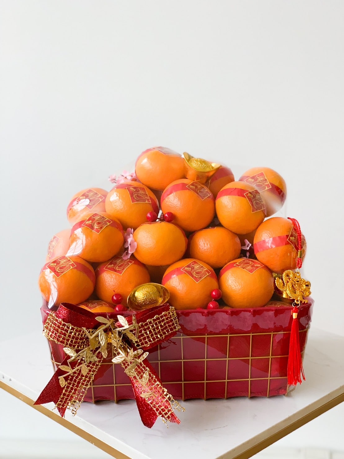 A red and gold basket filled with neatly arranged oranges, adorned with red ribbons, gold decorations, and festive charms. The basket sits on a white surface against a plain background, reflecting a celebratory and vibrant feel.