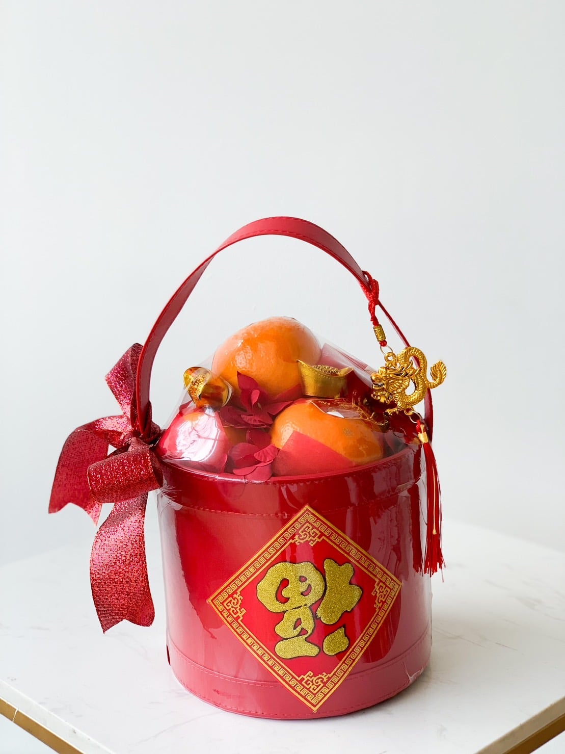 A round red basket with a red handle and bow, filled with oranges and a decorative gold charm. The basket features a yellow Chinese character on the front and is placed on a white surface. The background is plain white.