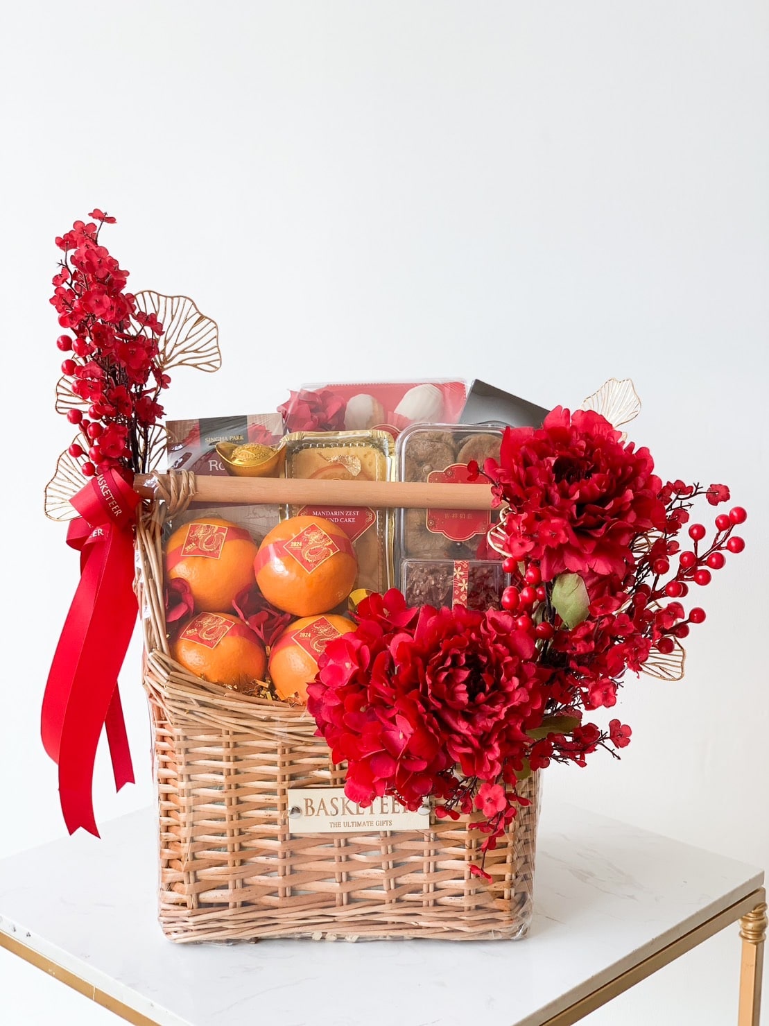 A wicker basket adorned with red flowers and ribbons contains an assortment of gifts. The basket includes oranges, boxes of candies or snacks, and decorative elements, making it a festive presentation. The basket is placed on a white surface against a plain background.