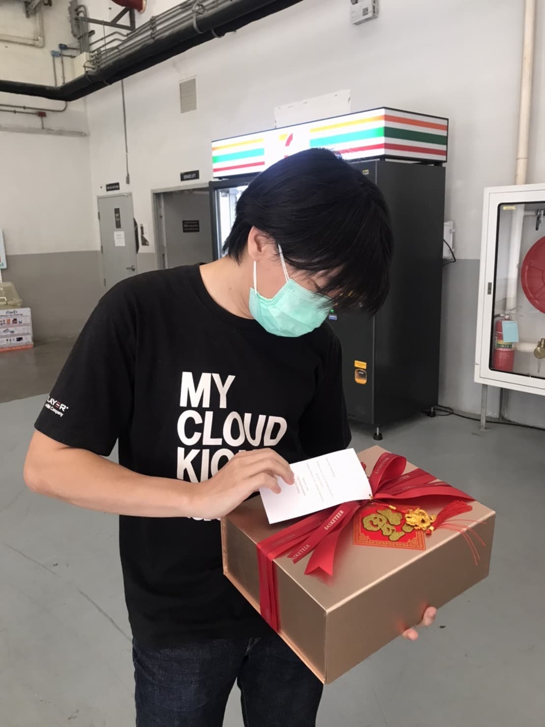 A person wearing a black t-shirt and a surgical mask is holding a wrapped gift with a red ribbon and decorative stickers. They appear to be reading a card attached to the gift. The background shows industrial equipment and a vending machine.