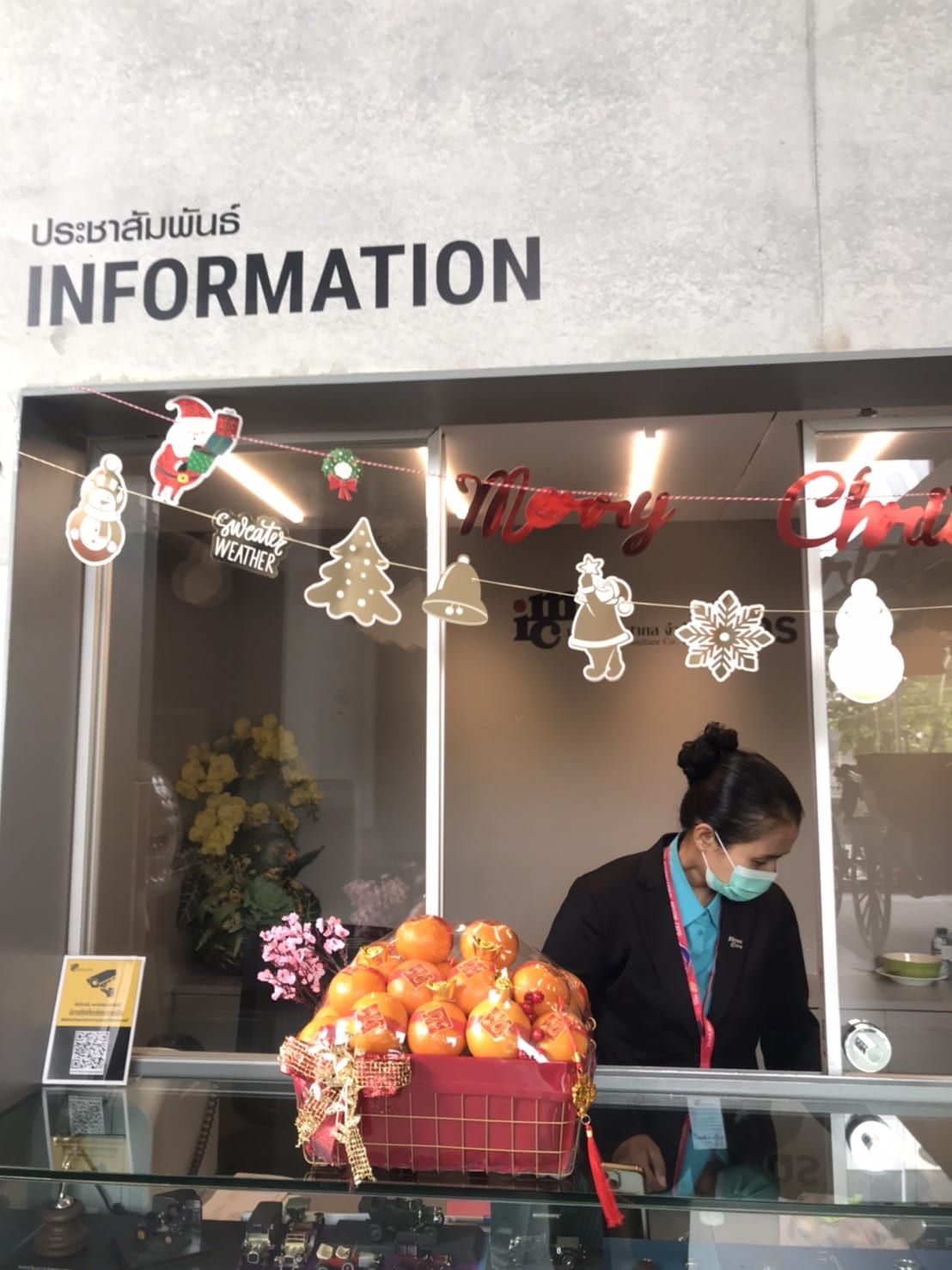 A woman wearing a mask and a black blazer stands behind an information counter decorated with Christmas ornaments and signs. In front of the counter is a basket filled with gift-wrapped boxes and orange items. The sign above the counter reads 