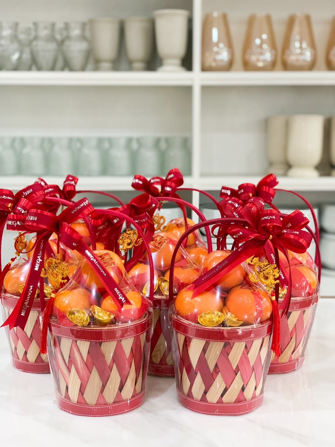 Six Majestic Wealth Mandarin Orange Hampers filled with oranges and gold-wrapped coins, each adorned with a red ribbon and bow, are arranged on a white marble surface. In the background, shelves display various glass and ceramic containers.