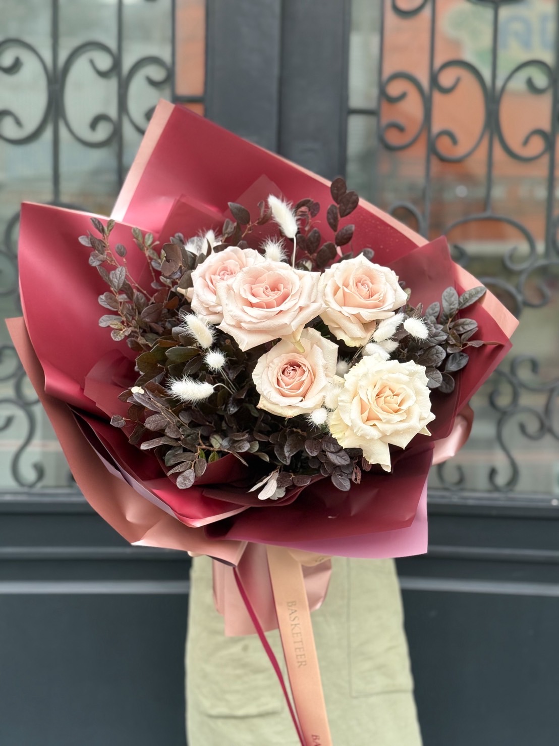 Elegant bouquet with blush pink and cream roses, accented with dark foliage and white dried flowers, wrapped in wine-red paper with Basketeer ribbon.