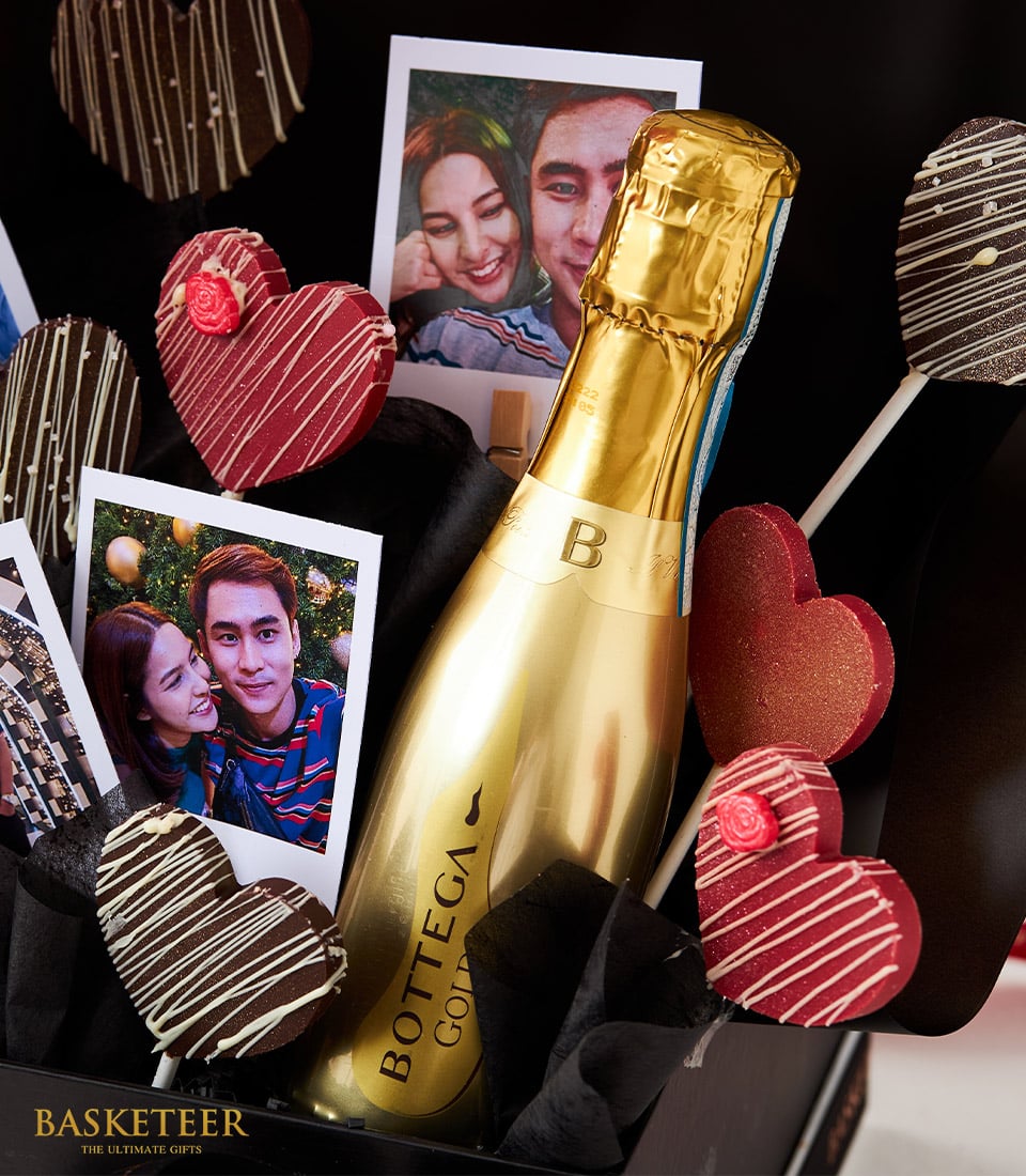 A luxurious gift basket featuring an elegant gold-tone bottle of Bottega prosecco, lavishly surrounded by decadent heart-shaped chocolate-coated lollipops and a vibrant heart balloon. Two beautifully printed photographs of a smiling couple add a personal touch to this exquisite scene. The prestigious Basketeer logo is artfully displayed at the bottom left corner, enhancing the sophistication and charm of this indulgent presentation.