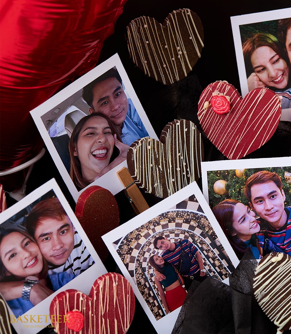 A captivating display showcasing a collection of Polaroid-style photos capturing the joyous smiles of a couple, elegantly interspersed with gourmet heart-shaped chocolates adorned with intricate white and red patterns. Set against a luxurious dark backdrop enriched with opulent red and black accents, the scene is further enhanced by an exquisite heart balloon that amplifies the romantic atmosphere. Basketeer branding is subtly present, evoking a sense of sophistication and exclusivity in this enchanting arrangement.