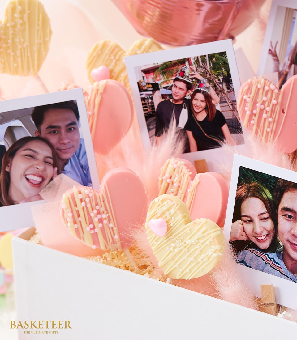 A luxurious gift arrangement showcases an assortment of heart-shaped cookies, elegantly adorned with delicate pink and white icing. Interspersed among the sweet treats are cherished printed photos of a smiling couple, capturing moments of joy and love. An enchanting pink heart balloon adds a whimsical flair, hovering above soft, inviting pink fluffy material within a pristine white container. The scene is beautifully completed with the iconic 
