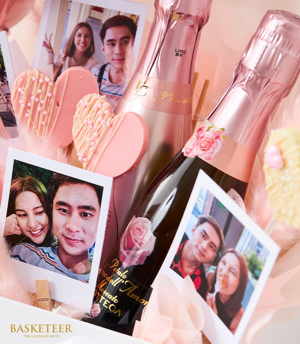 An opulent gift basket elegantly showcasing two bottles of exquisite sweet wine, their pink caps gleaming invitingly. Nestled beside them are two decadent heart-shaped pink cookies, crafted to perfection. Three pinned photographs capture the radiant smiles of a joyous couple, adding a personal touch. The basket is beautifully adorned with soft pink accents and features an enchanting pink heart balloon floating above. A sophisticated ribbon and box prominently display the Basketeer logo, encapsulating the brand’s essence - 