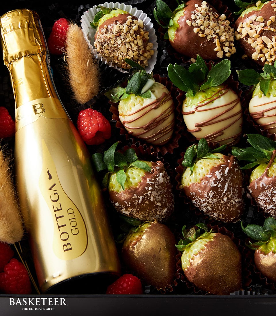 A luxurious close-up of an elegant gift arrangement by Basketeer. At its center is a refined bottle of Bottega Gold Prosecco, gleaming with sophistication. Surrounding it, an indulgent selection of chocolate-covered strawberries - some elegantly draped in white chocolate and adorned with a dusting of nuts or delicate coconut flakes. Scattered among them are vibrant red raspberries and exquisite decorative elements, creating an inviting scene that exudes opulence and taste. The Basketeer logo subtly graces a ribbon or box within the composition, signifying quality and luxury at every detail.