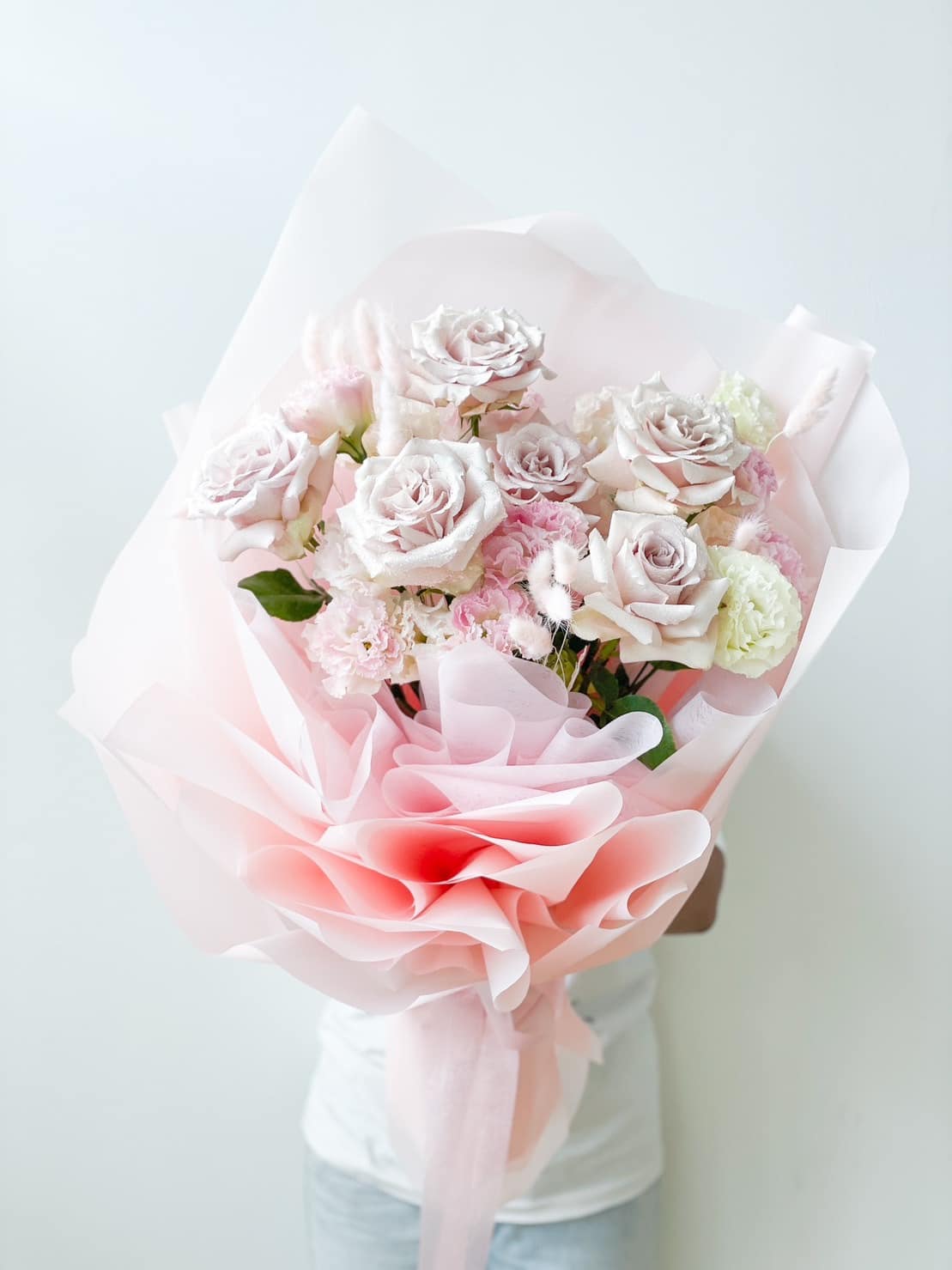 A person holds a large bouquet of pale pink and cream roses wrapped in light pink and white paper. The bouquet features different sizes of roses and greenery, creating a soft, romantic arrangement against a plain white background.