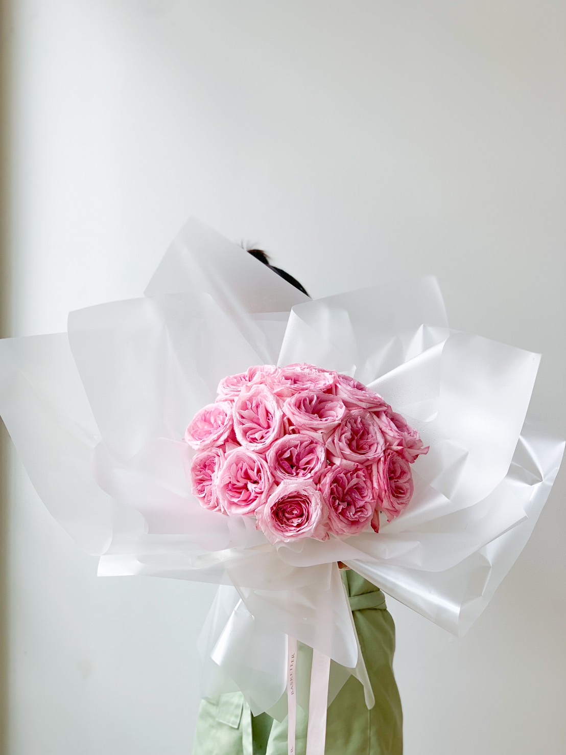 A person holds a sweet pink roses bouquet wrapped in white translucent paper. The large bouquet obscures the person's face, and they are wearing a light green top. The background is a plain, light-colored wall.