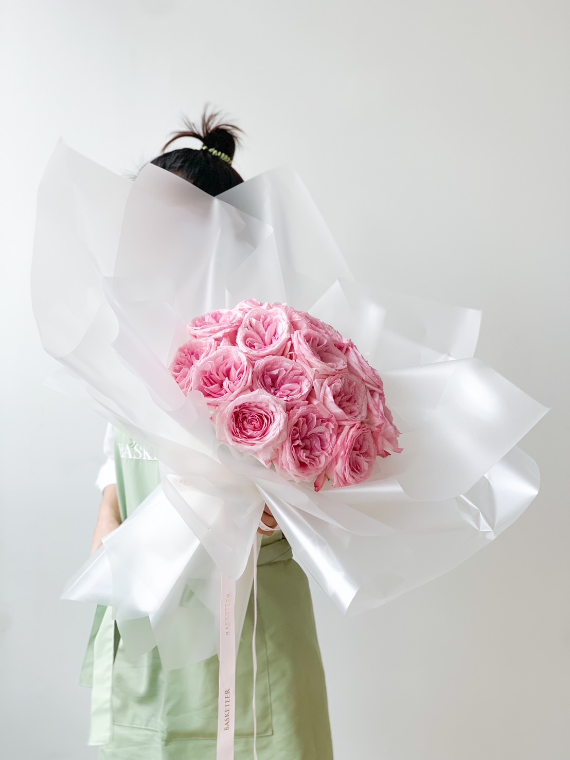 A person in a green apron holds a large Sweet Pink Roses Bouquet wrapped in translucent white paper against a plain white background. The bouquet is voluminous, with roses in full bloom, and the individual’s face is partially obscured by the flowers.