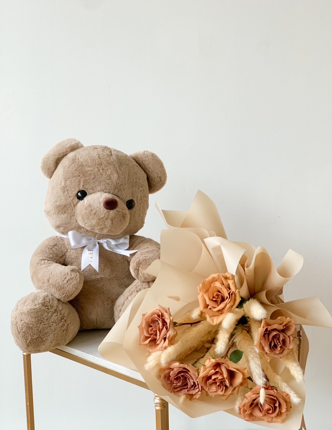 A cuddly brown teddy bear with a white bow sits on a small table next to a Brown Teddy Bear Toffee Bouquet Set, featuring peach-colored roses wrapped in light beige paper. The background is a plain white wall.