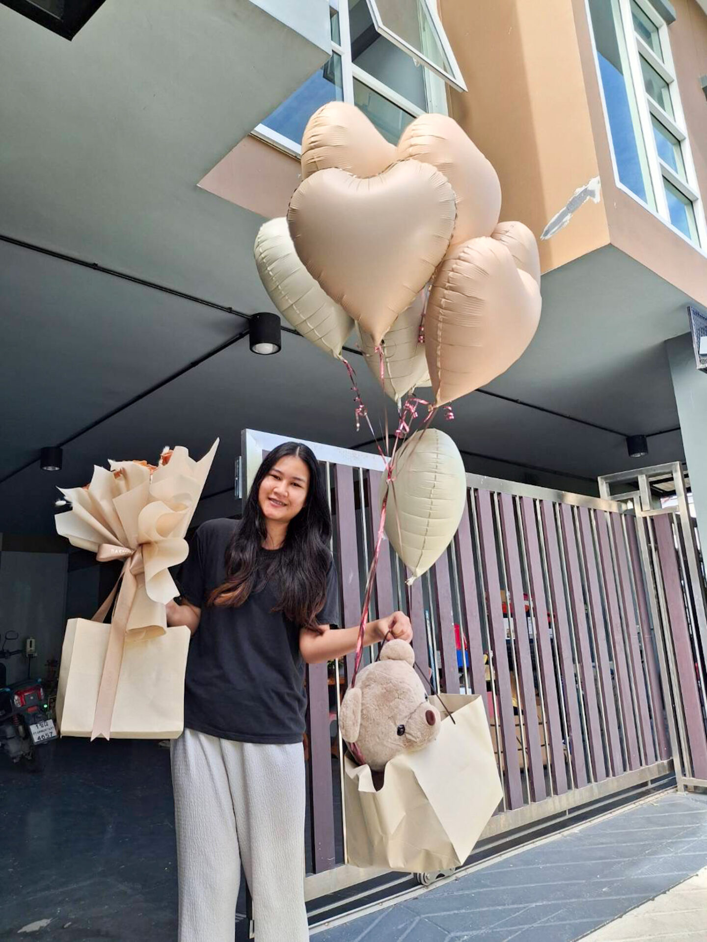 A woman stands outdoors holding heart-shaped balloons and a toffee bouquet set in one hand, while clutching gift bags and a brown teddy bear in the other. She is smiling and dressed casually in a black shirt and gray pants, standing in front of a modern building with large windows and a gated entrance.
