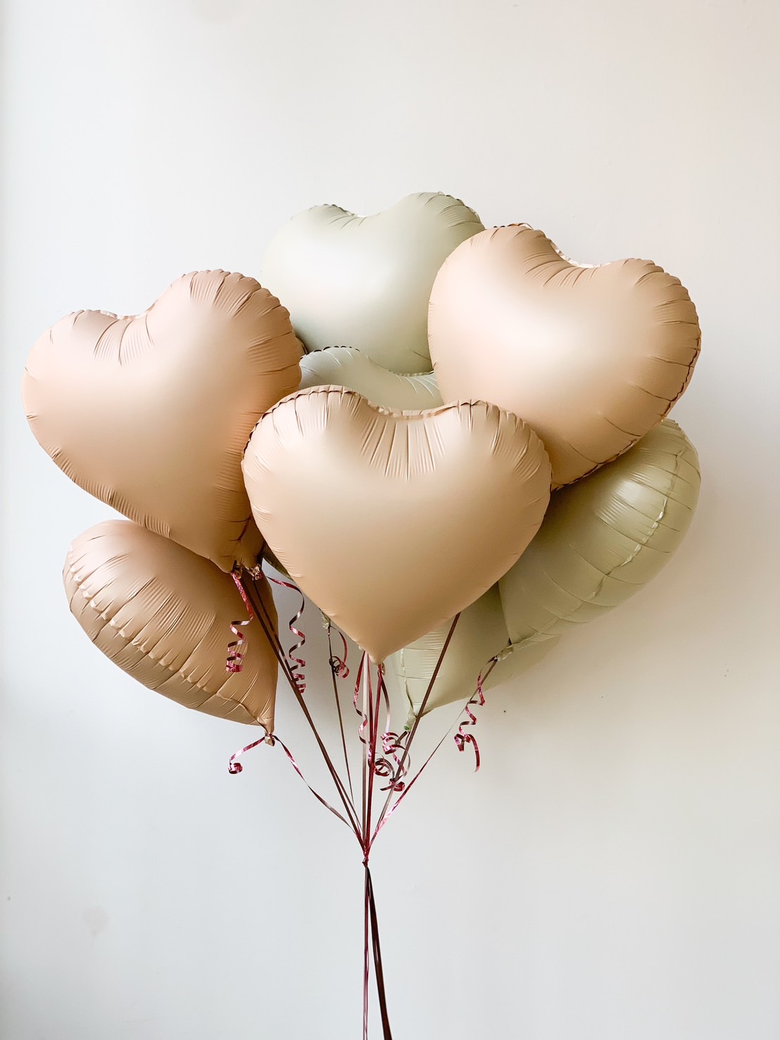 A bunch of heart-shaped balloons in soft pastel shades of beige and light green float gracefully, attached to strings against a plain, light-colored background. Beside them, a charming brown teddy bear holds a delightful toffee bouquet set, creating a sweet and endearing scene.