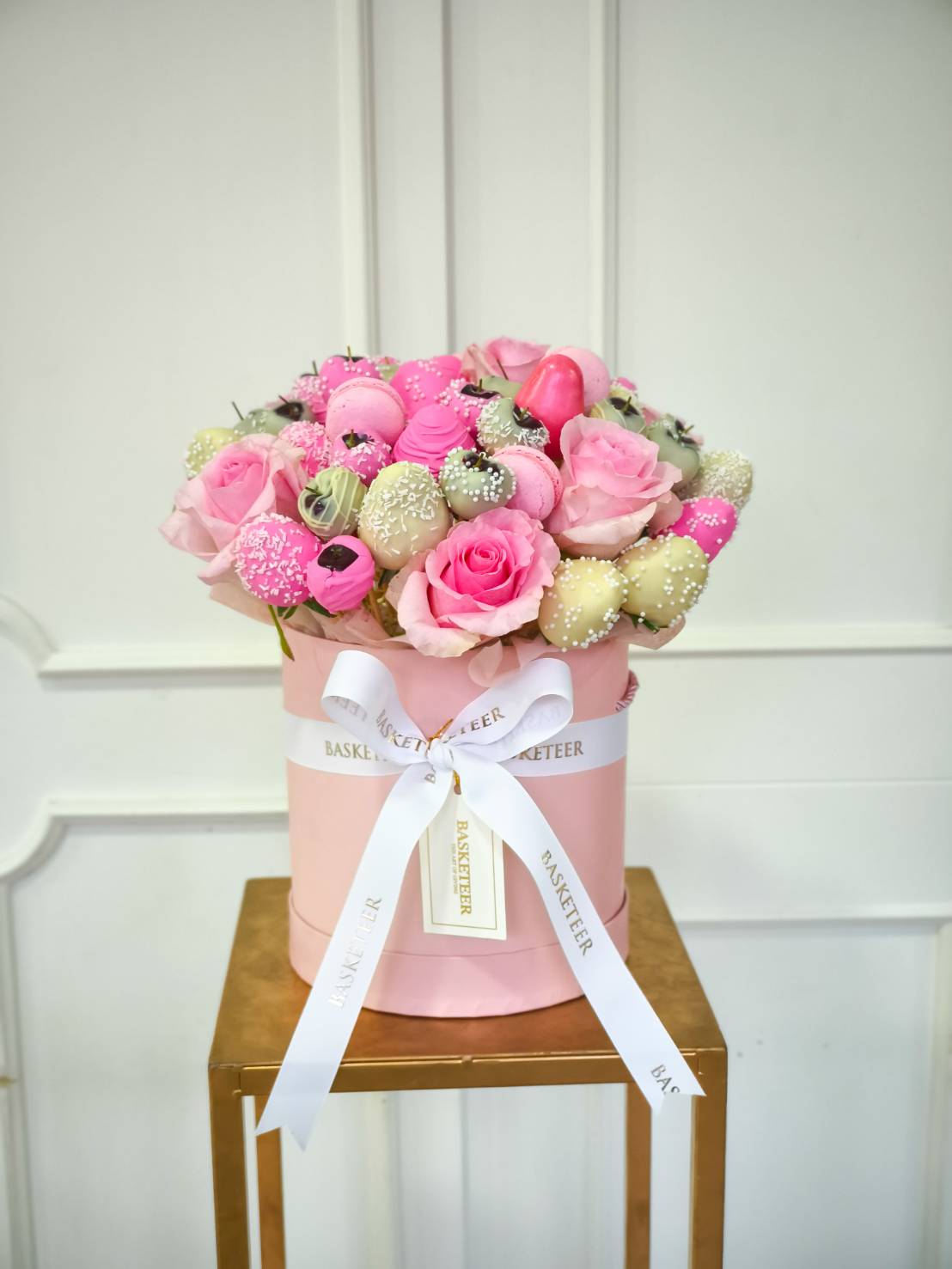 Pink hatbox arrangement with pink roses, macarons, and white chocolate-covered strawberries decorated with sprinkles, tied with a white Basketeer ribbon.