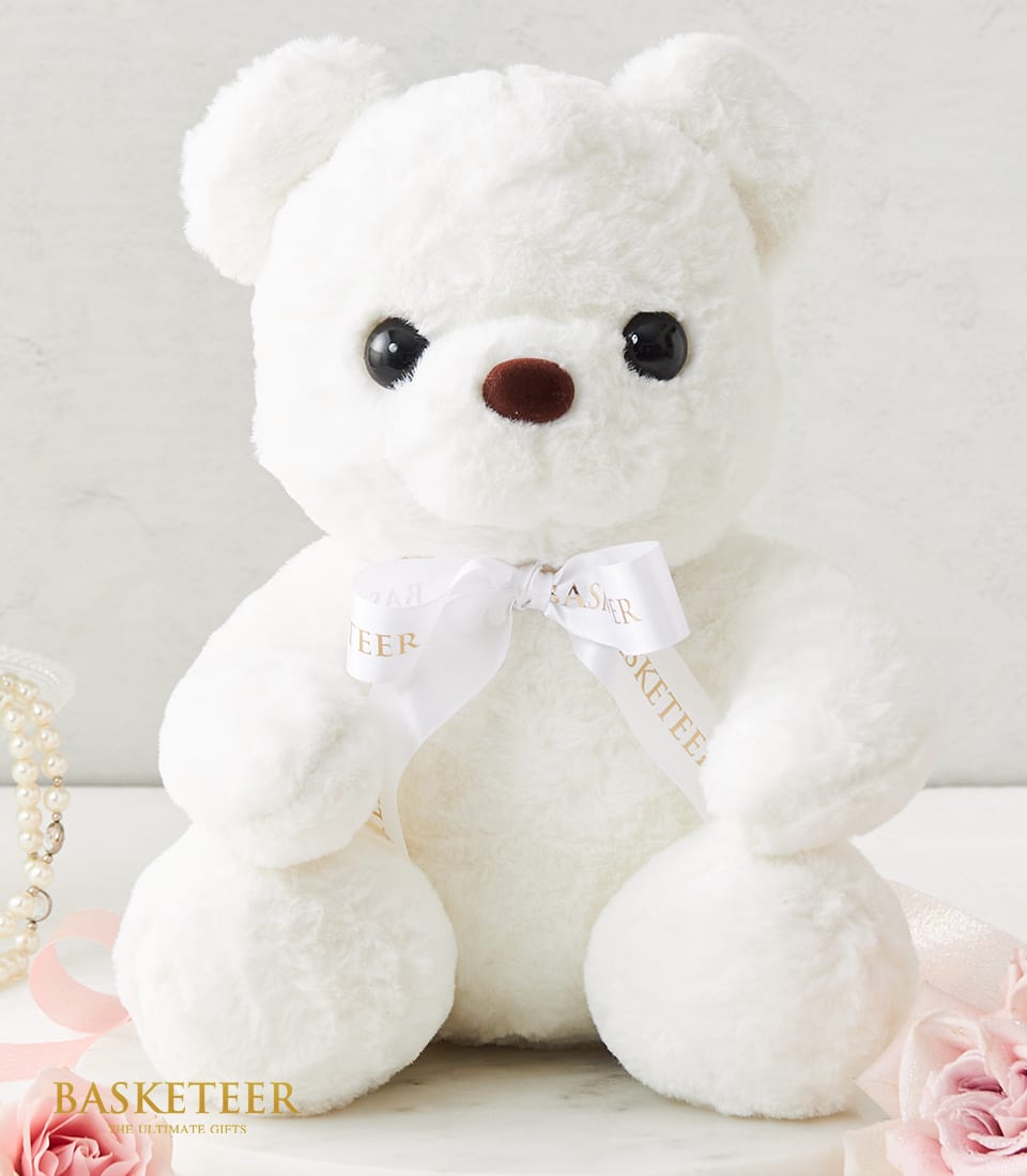 A plush white teddy bear with soulful dark eyes and a charming brown nose is gracefully adorned with an elegant white ribbon that proudly displays 