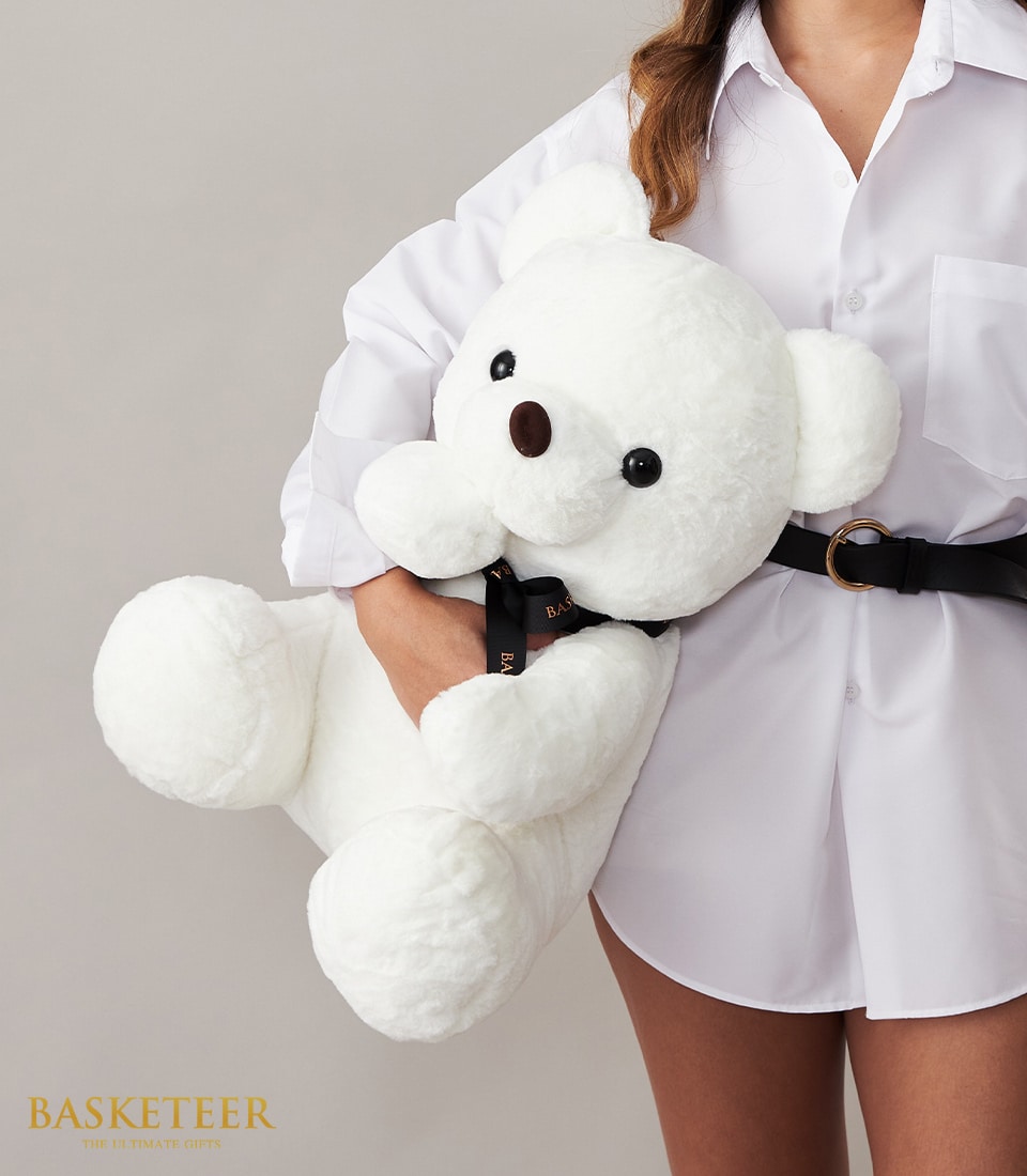 A person elegantly adorned in a crisp white shirt delicately cradles a sumptuous, oversized plush teddy bear. This luxurious teddy bear boasts a charming brown nose, soulful black eyes, and is adorned with an opulent black bow featuring gold text. The serene backdrop highlights the sophisticated simplicity of the scene. In the bottom left corner, the distinguished 