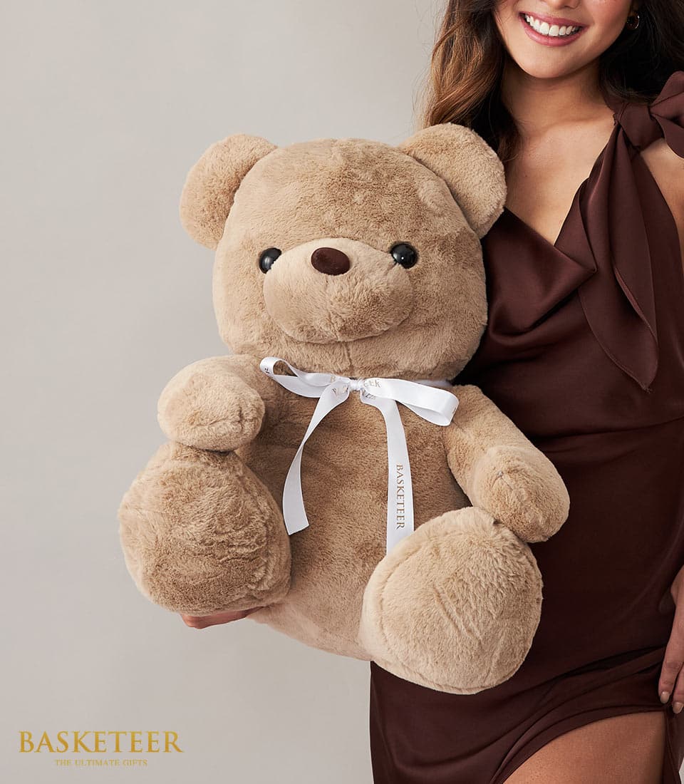 A joyful woman in an elegant brown dress beams as she embraces a luxurious, oversized light brown teddy bear, which is beautifully adorned with a white ribbon elegantly inscribed with 