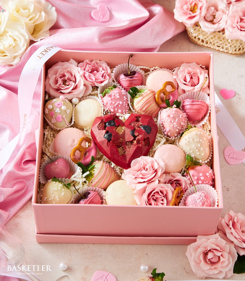 A luxurious pink gift box elegantly adorned with Basketeer branding, filled with an exquisite assortment of confections. Inside, discover a decadent red heart-shaped candy, delicate pink macarons, chocolate-dipped pretzels, a variety of gourmet chocolates, and pastel pink roses. The box is beautifully decorated with sumptuous pink and white ribbons bearing the Basketeer logo. The scene is completed with lush pink roses and opulent fabrics that evoke an inviting sense of elegance and refinement.