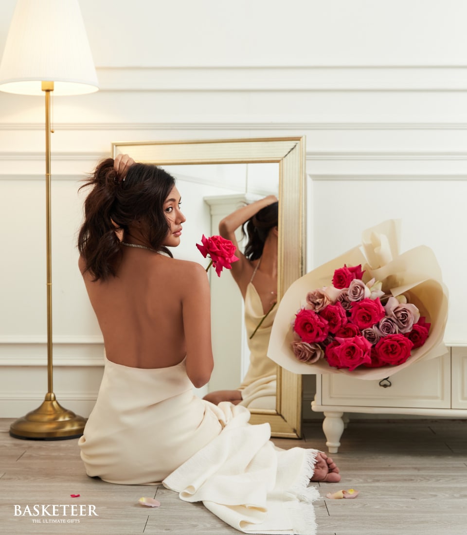 A woman with a graceful, bare back sits on an opulent floor in front of an exquisite mirror, delicately holding lush red roses near her face. Beside her, on a pristine white table adorned with the Basketeer logo, lies a sumptuous bouquet of red and pink roses, radiating a romantic charm. As she gently touches her flowing hair with one hand, an elegant lamp casts a soft glow over the sophisticated and minimalist setting. This serene moment captures the luxury and inviting allure that Basketeer brings to your most cherished spaces.