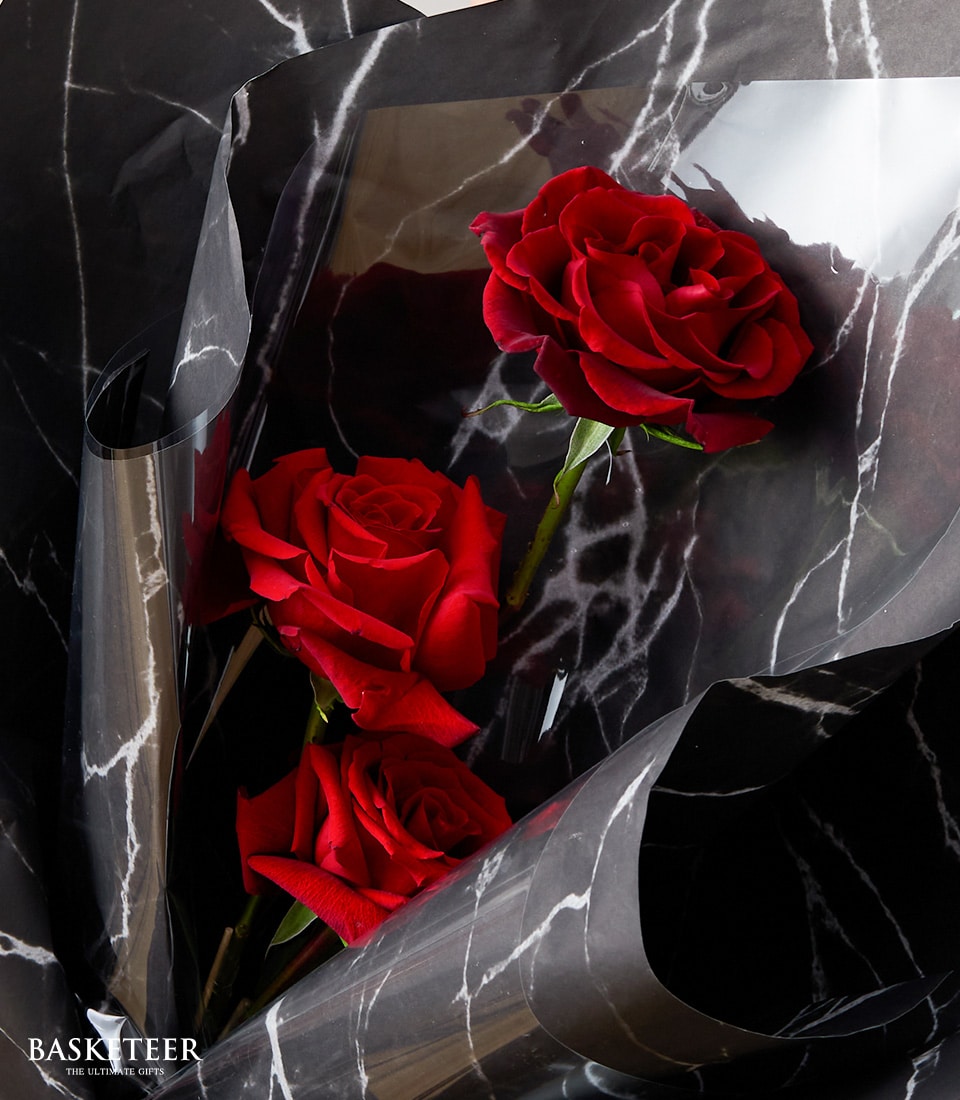 A luxurious bouquet of three vibrant red roses elegantly wrapped in sophisticated black and white marble-patterned paper. The delicate petals exude opulence, while the rich green stems offer a striking contrast against the sleek backdrop. In the bottom left corner, the distinguished Basketeer logo adds a touch of refinement, epitomizing upscale elegance and inviting allure.