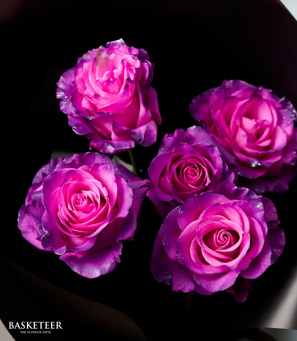 A lavish bouquet of five exquisite pink and purple roses with glittering edges, elegantly arranged in a circular formation against an opulent dark backdrop. A sophisticated branded label displaying 