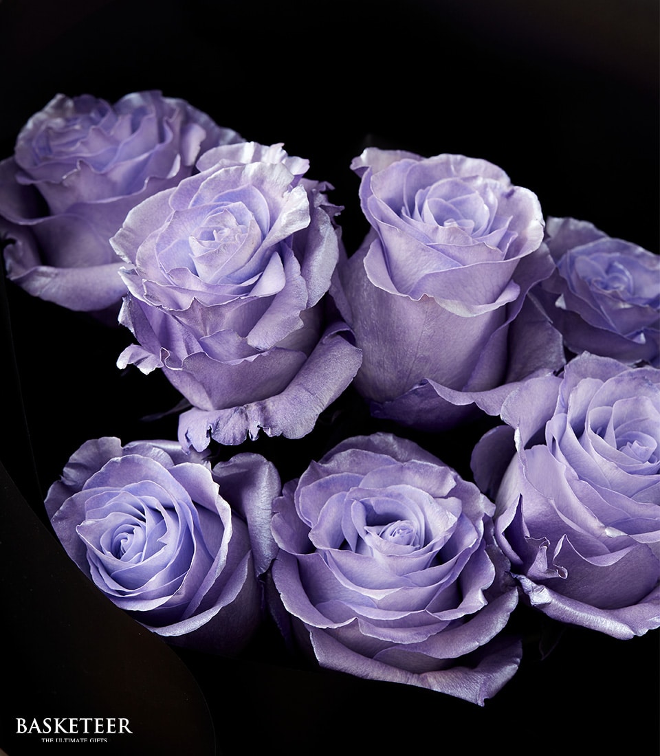 A close-up of six exquisite lavender-colored roses, meticulously arranged against an opulent dark background. The petals are intricately layered, revealing deep and luxurious violet tones. In the bottom left corner, a sophisticated watermark reads 