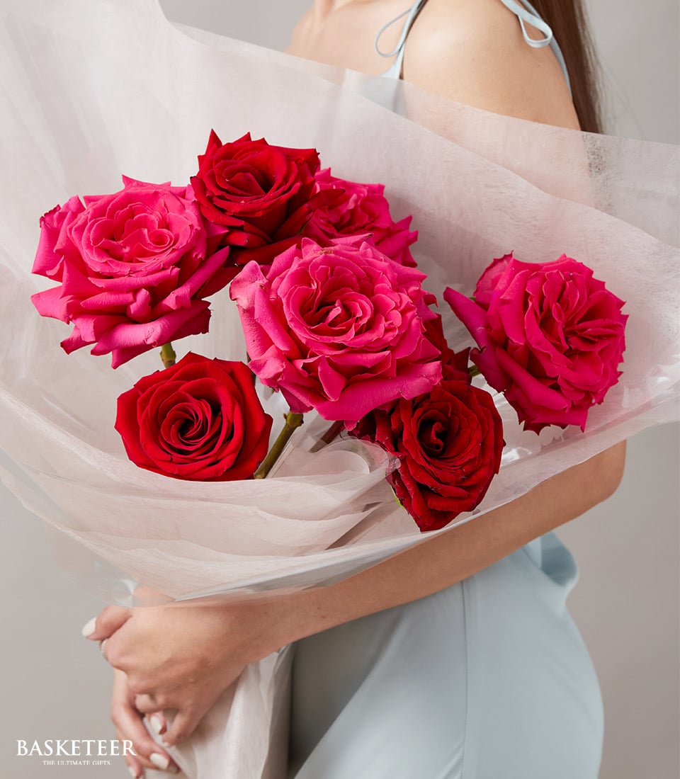 A person in an elegant light blue dress cradles a sumptuous bouquet of vibrant red and pink roses, exquisitely wrapped in pristine white tissue paper. The serene gray backdrop accentuates the richness of the blooms, evoking a sense of timeless luxury. At the bottom left corner, the sophisticated Basketeer logo and 